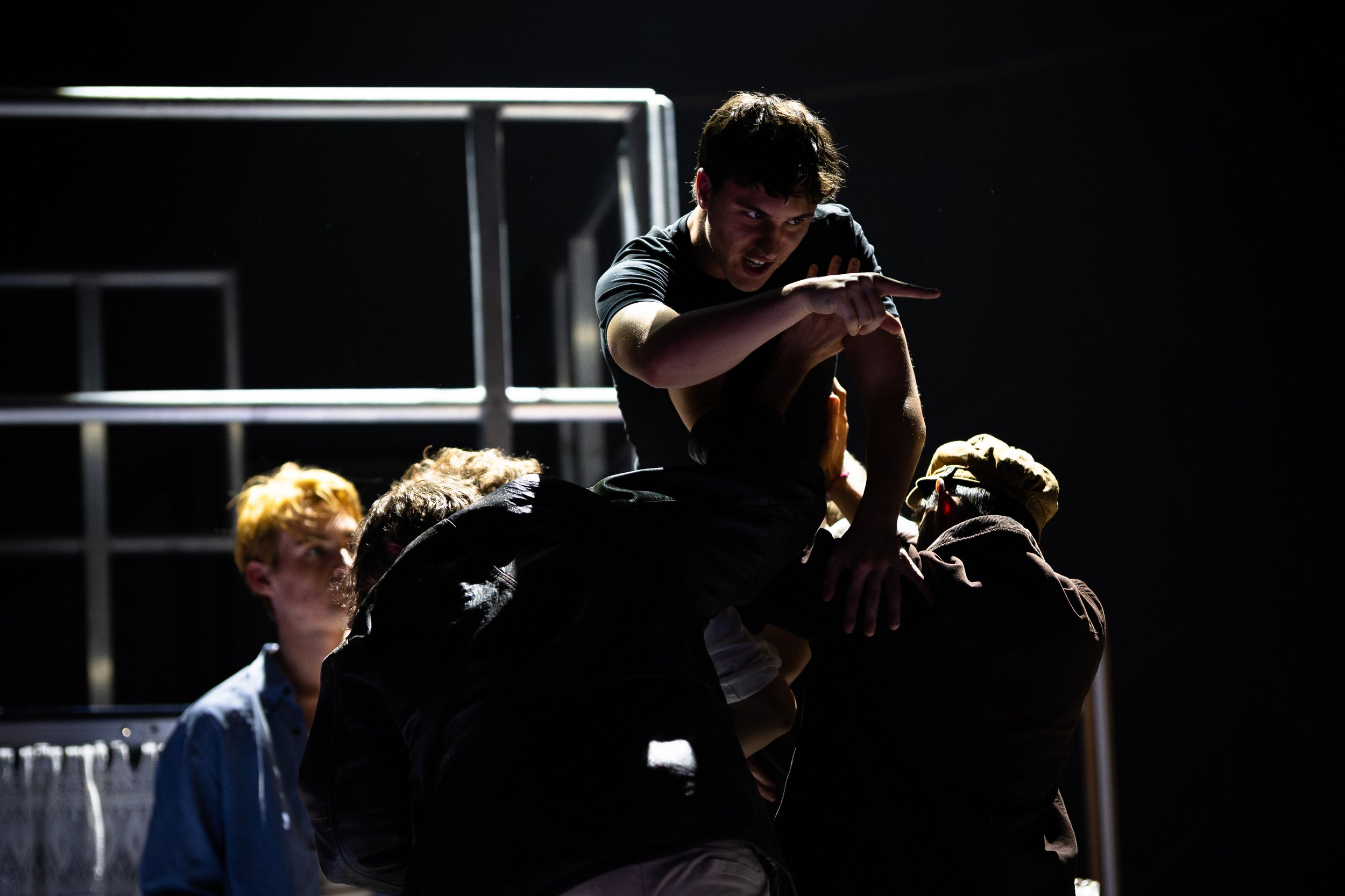 Group of people dressed in dark clothing performing a coordinated human pyramid on a stage with minimal lighting.
