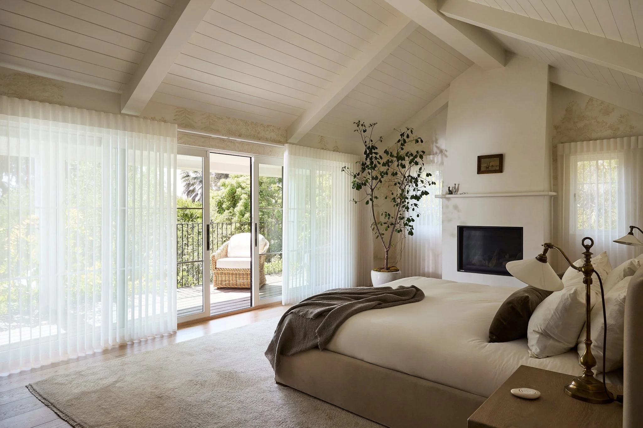 A cozy bedroom with white walls and ceiling, large windows with sheer white curtains, a balcony outdoor chair, a potted tree, a flat-screen TV on the wall, a bed with beige and brown bedding, a bedside table with a lamp, and natural light from the outside.