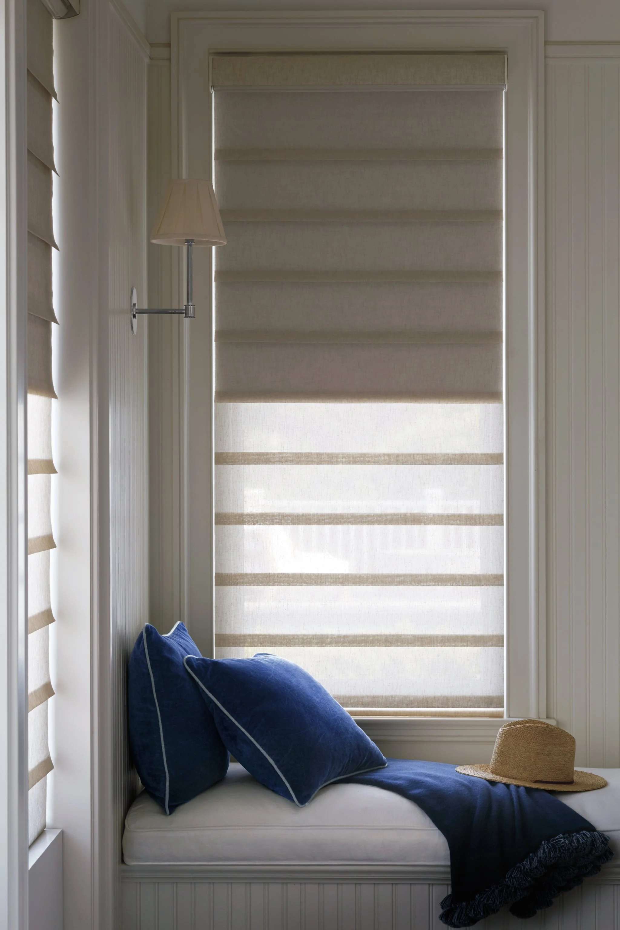 A window with beige striped blinds, a cushioned window seat with blue pillows and a straw hat, a wall-mounted lamp, and partially visible beige chairs in a cozy, well-lit room.