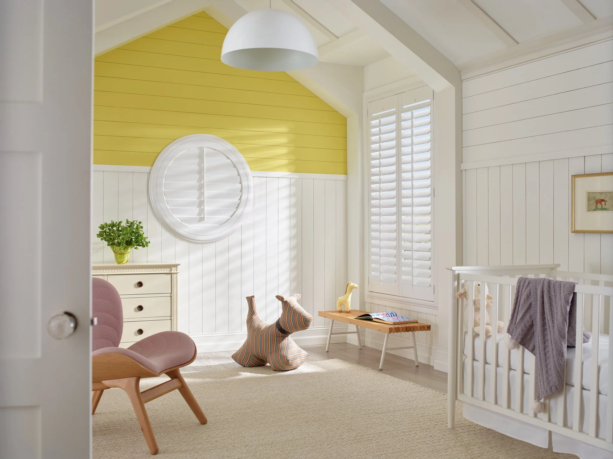 A cozy nursery with white wood-paneled walls, a yellow accent wall, a white ceiling with an angled design, and a window. The room features pink velvet chairs, a wooden bench, a colorful striped plush dog, framed wall art, a small potted plant