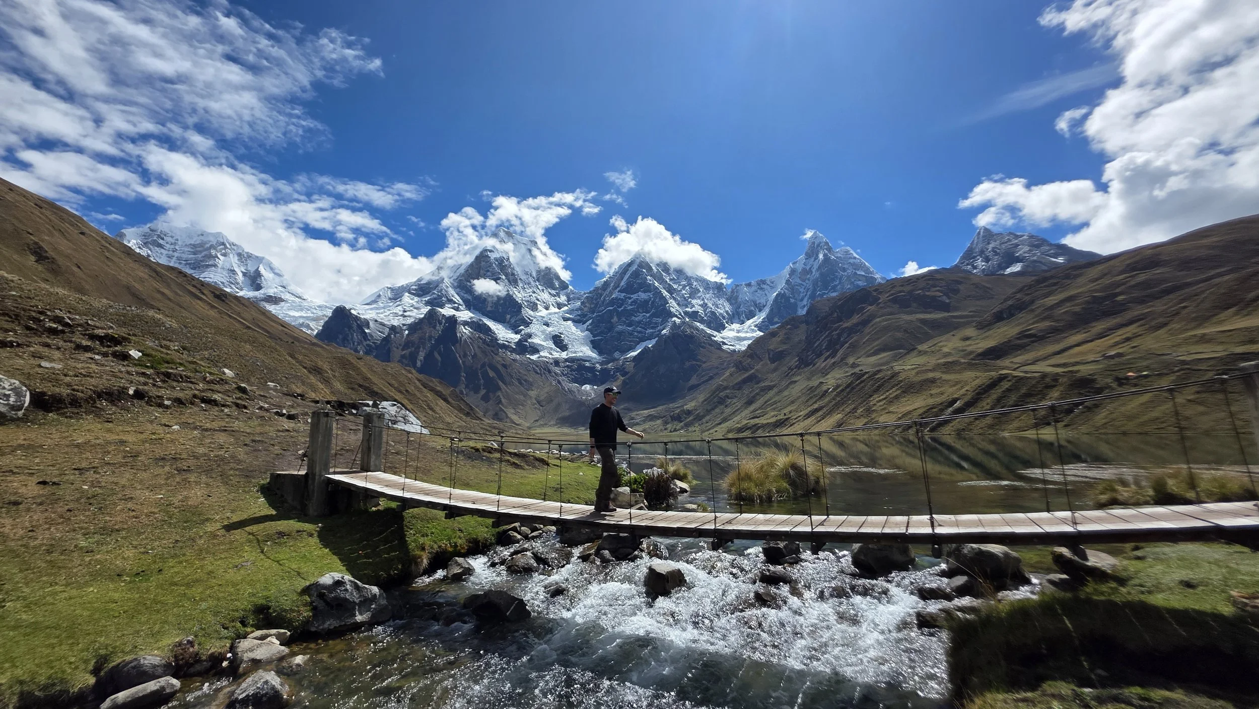 Person walking on a small wooden bridge over a stream in a mountainous area with snow-capped peaks, green hills, and a partly cloudy blue sky in the background.
