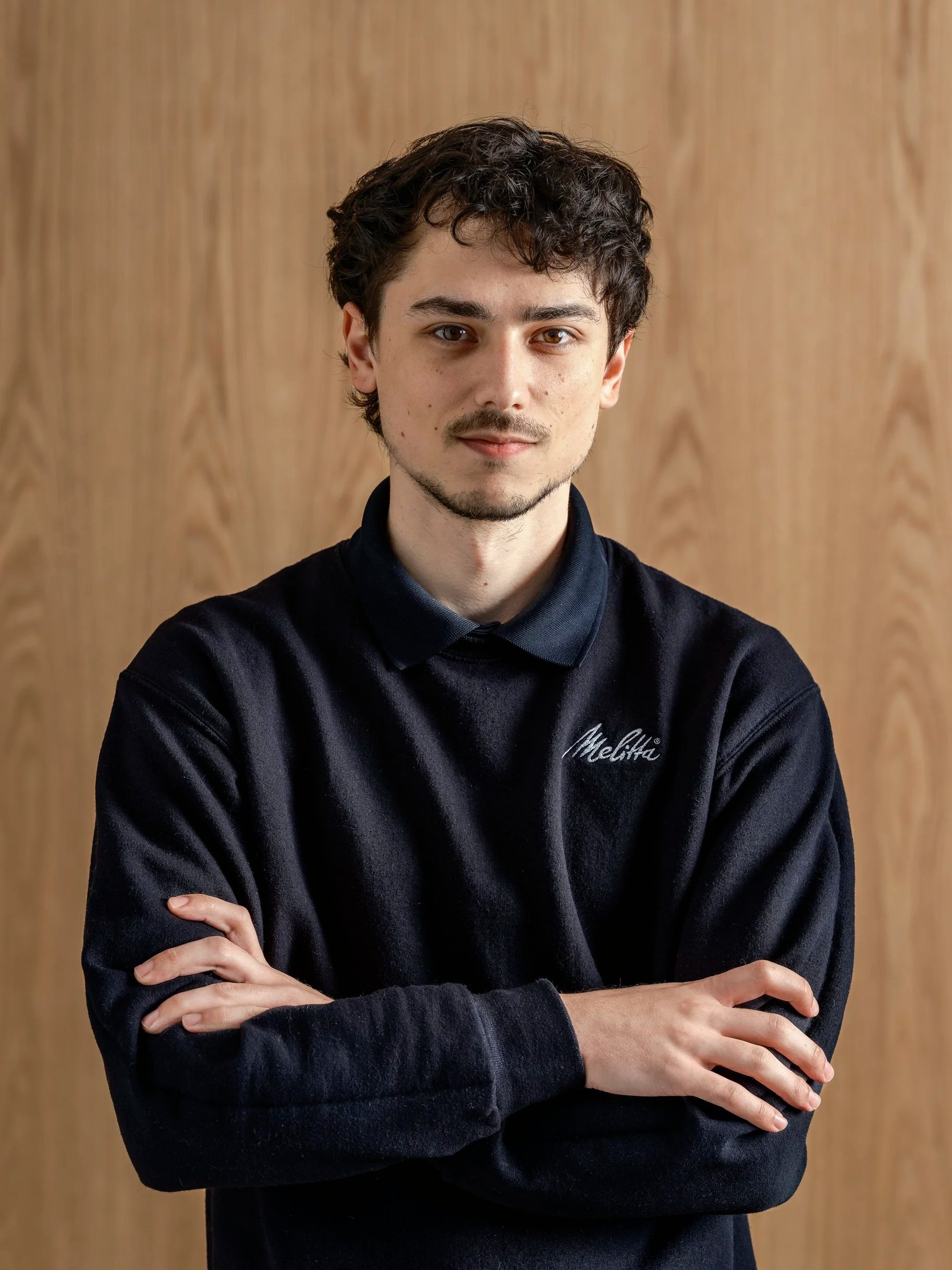 A young man with dark curly hair, wearing a black Melitta hoodie, standing with arms crossed in front of a wooden background.