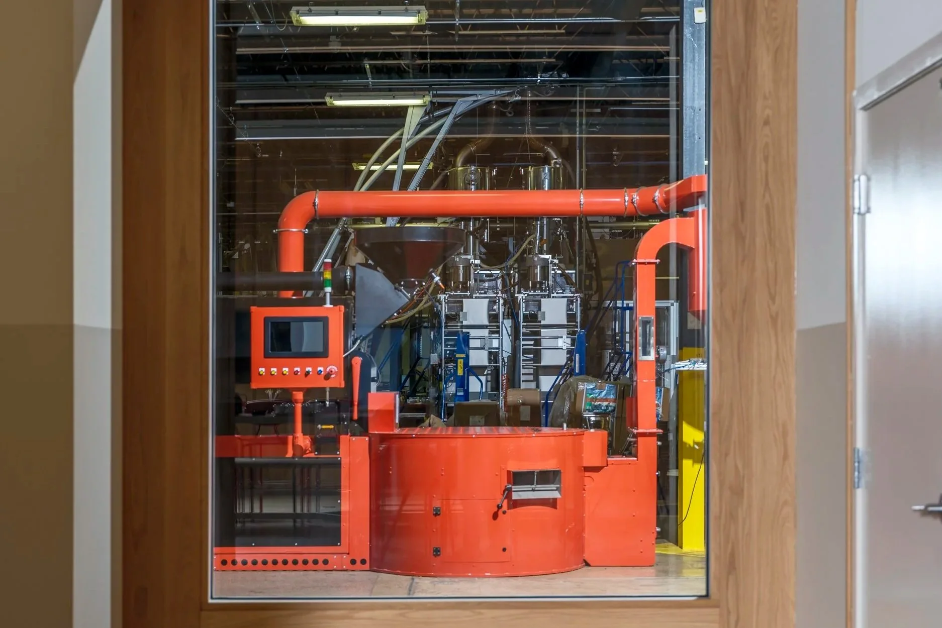 A large, industrial-looking machine behind a glass window, with various pipes, controls, and metal parts, situated in a room with wooden walls.