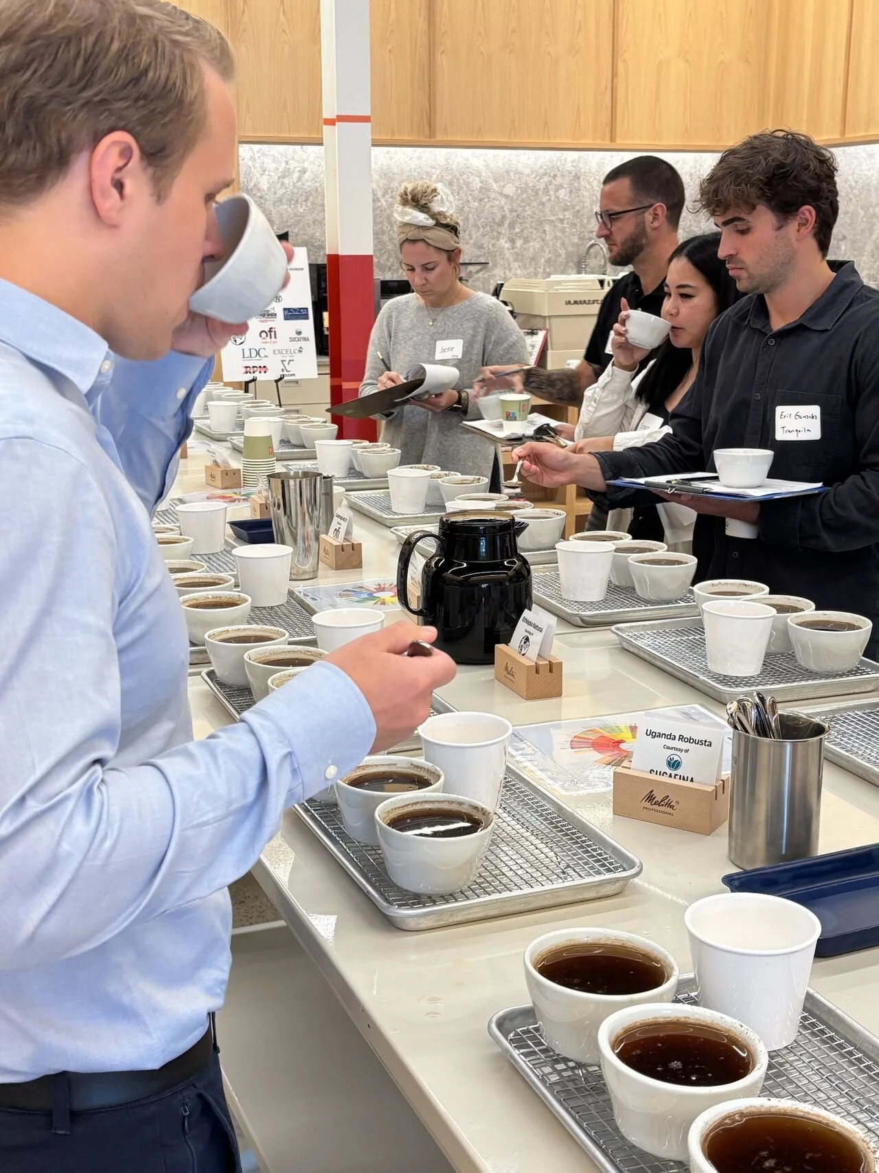 Group of six people in meeting room, two standing, four seated at table, coffee machines in background.