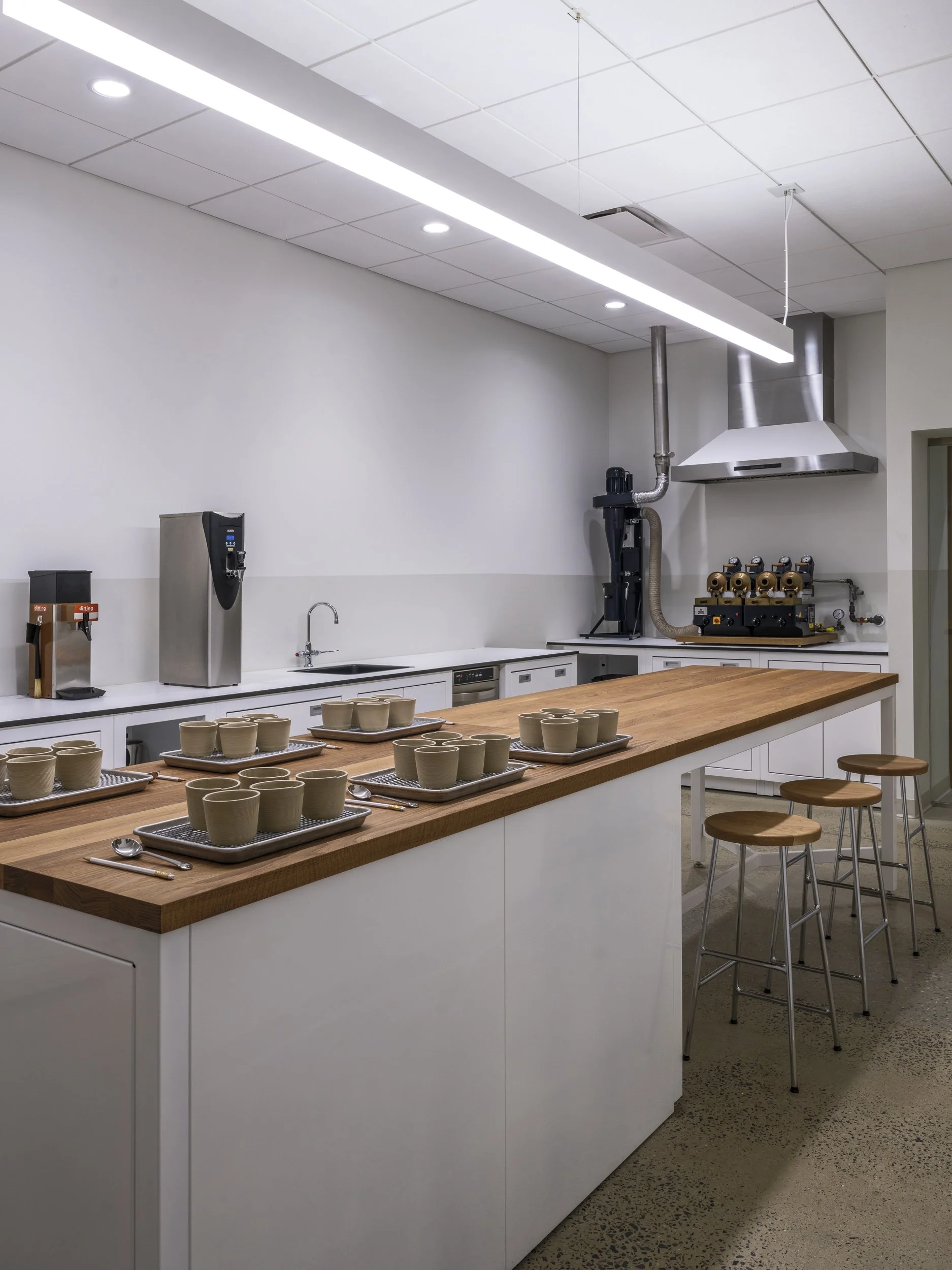 A modern kitchen or lab with a long wooden table set with numerous beige cups and small spoons, white cabinets, a water dispenser, and industrial equipment, under a white drop ceiling with embedded lighting.