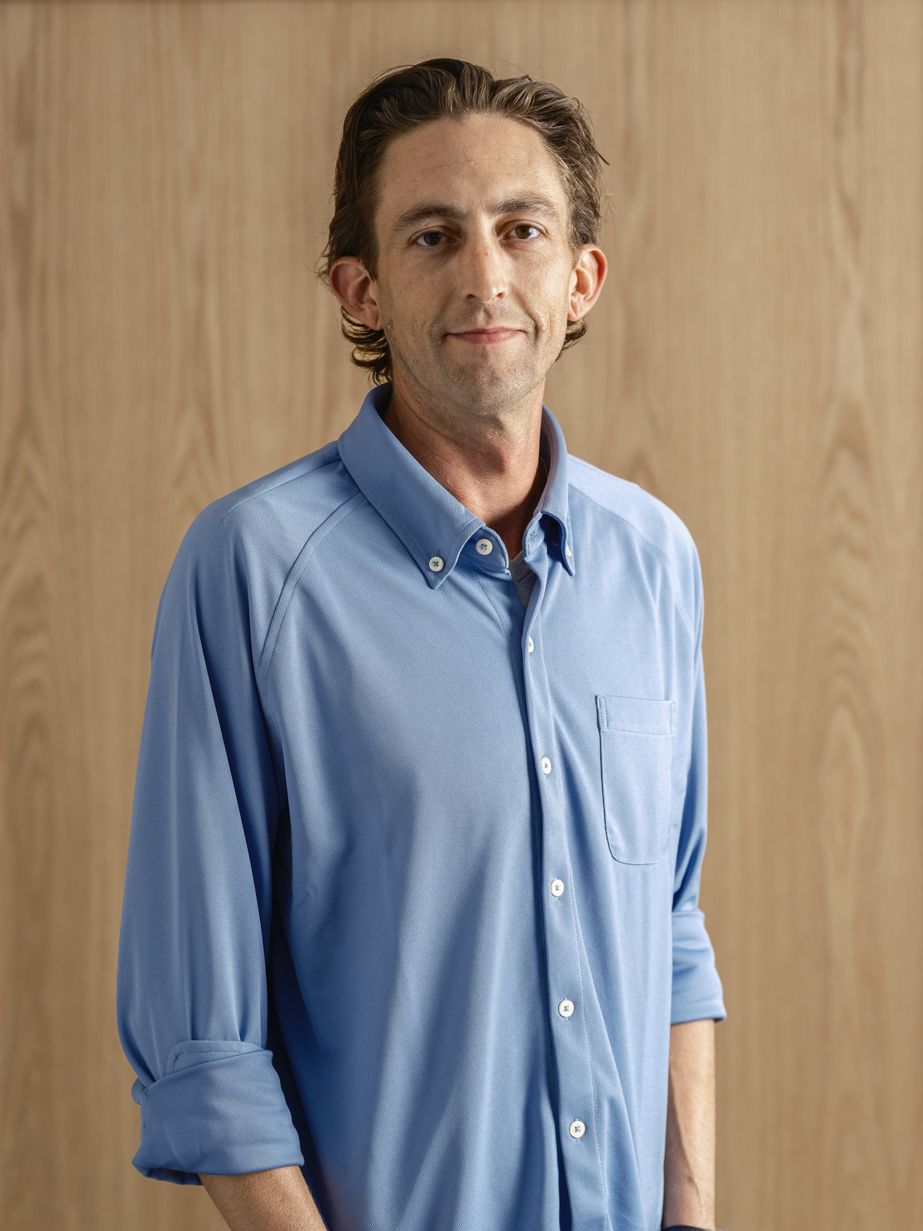 A man with light skin, brown hair, and a gentle smile wearing a light blue button-up shirt with rolled-up sleeves standing against a wooden background.