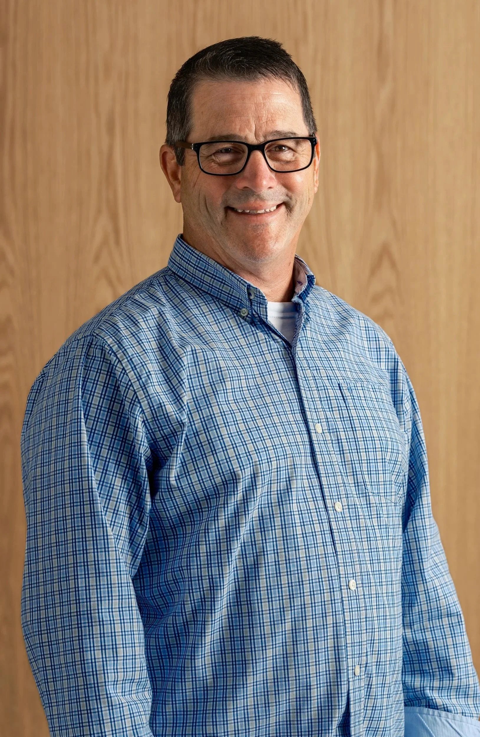A middle-aged man smiling, wearing black glasses, a blue checkered shirt, standing in front of a wooden background.