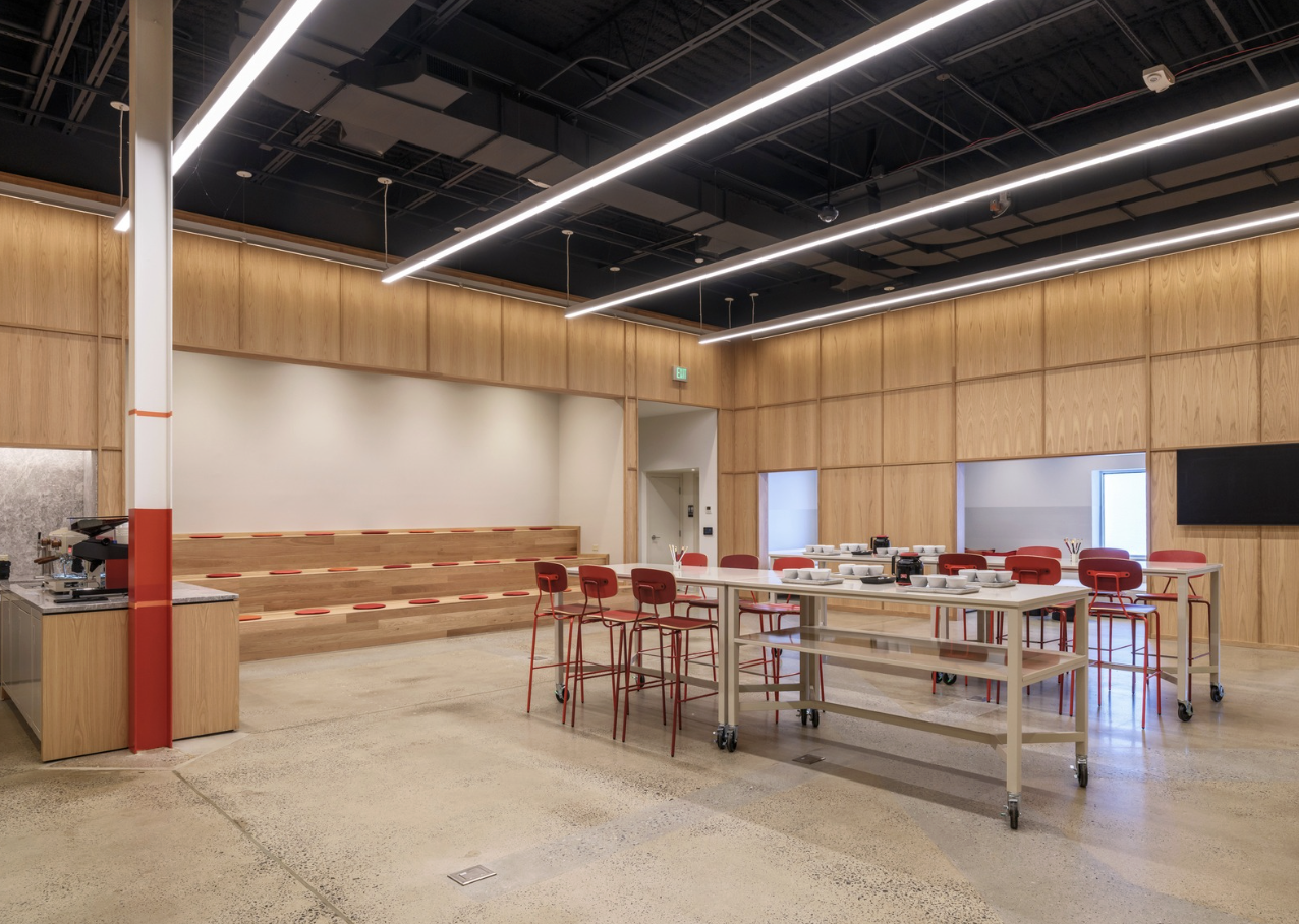 Modern conference room with tiered seating, red chairs, a long table with tableware, and a black wall-mounted monitor, featuring wood-paneled walls and high ceilings with linear lighting.
