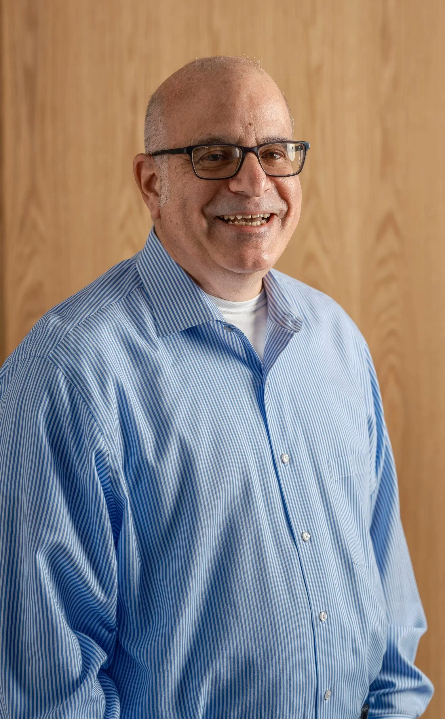 Portrait of a middle-aged man wearing glasses and a blue striped dress shirt, smiling against a light wooden background.