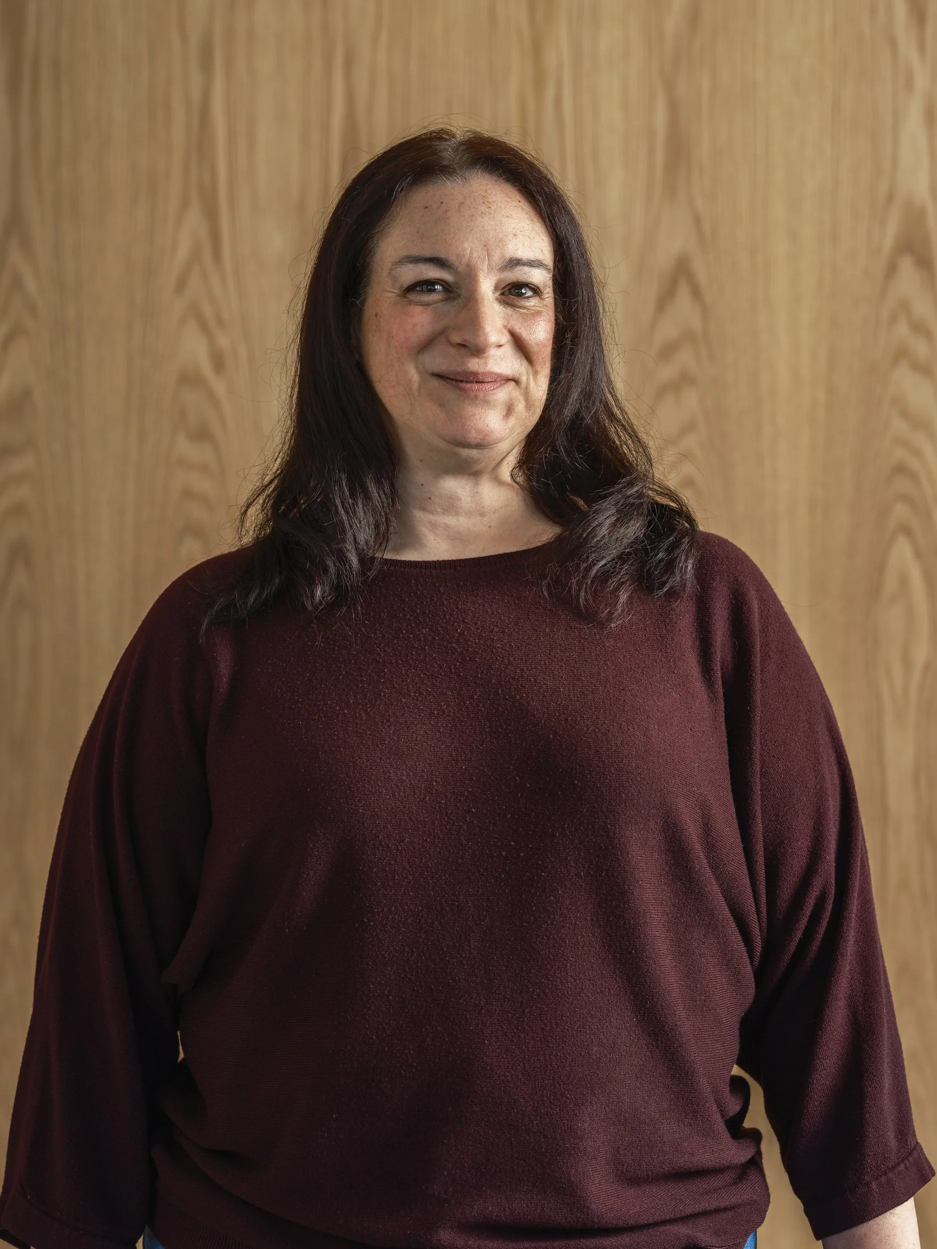 A woman with dark brown hair wearing a maroon dress standing against a wooden wall.