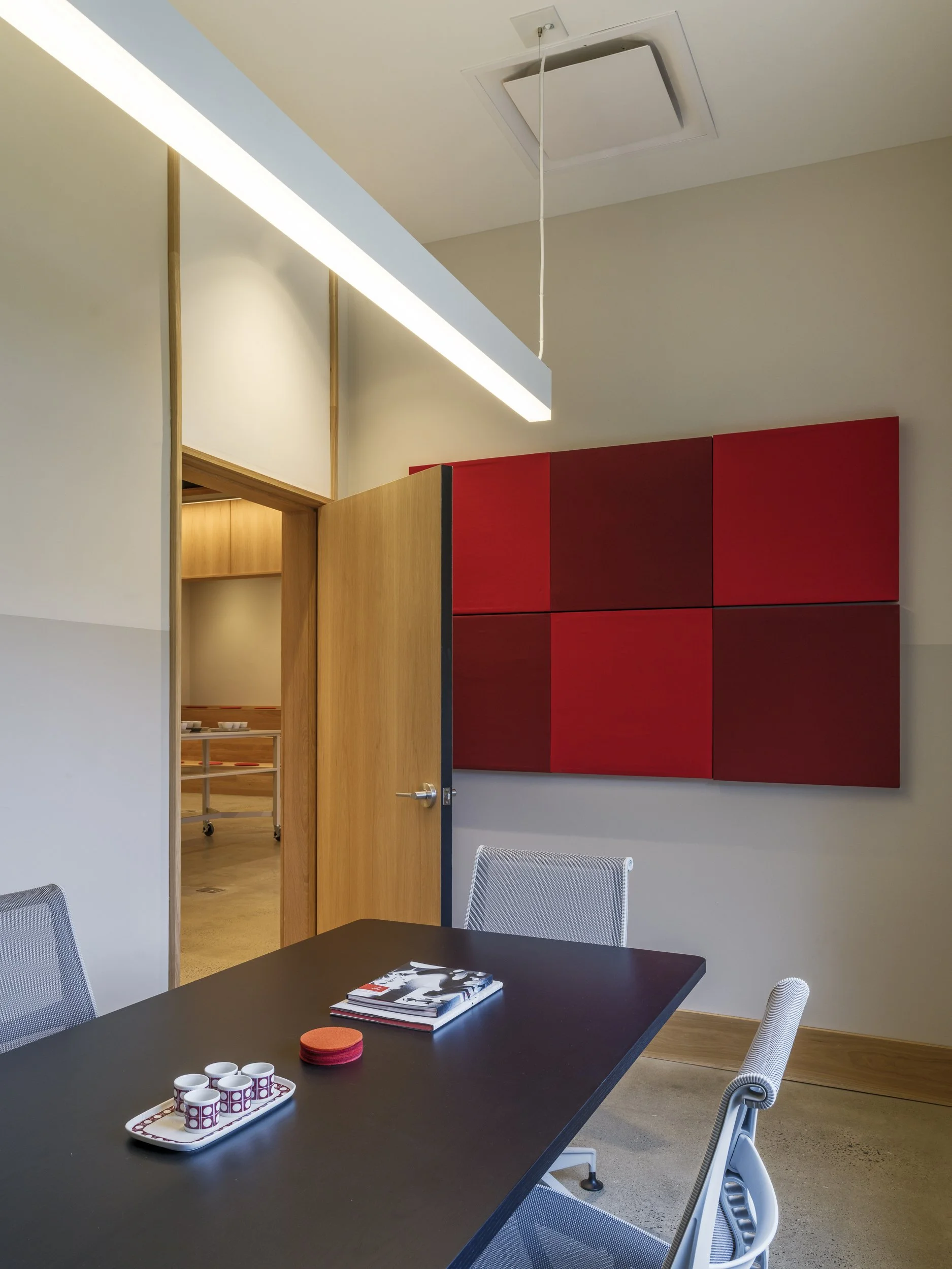 Meeting room with a black table, white chairs, a red and black wall art, and a doorway leading to a room with a table and chairs. There are some cups and a book on the table.