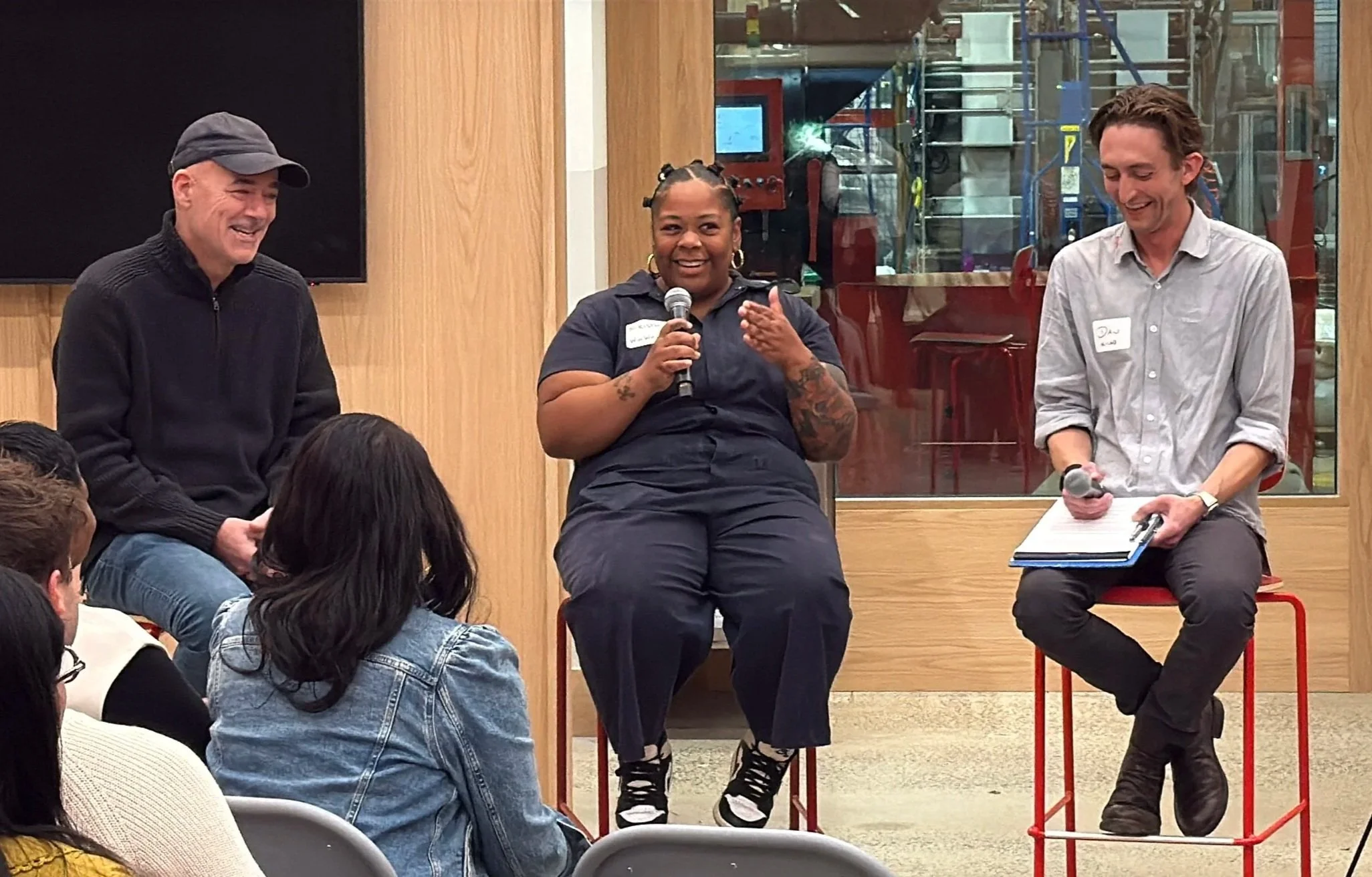Three people sitting on chairs, speaking or listening in a panel discussion inside a room with wooden walls and a glass window showing an industrial or factory setting in the background. Audience members are seated in front, with some visible in the foreground.
