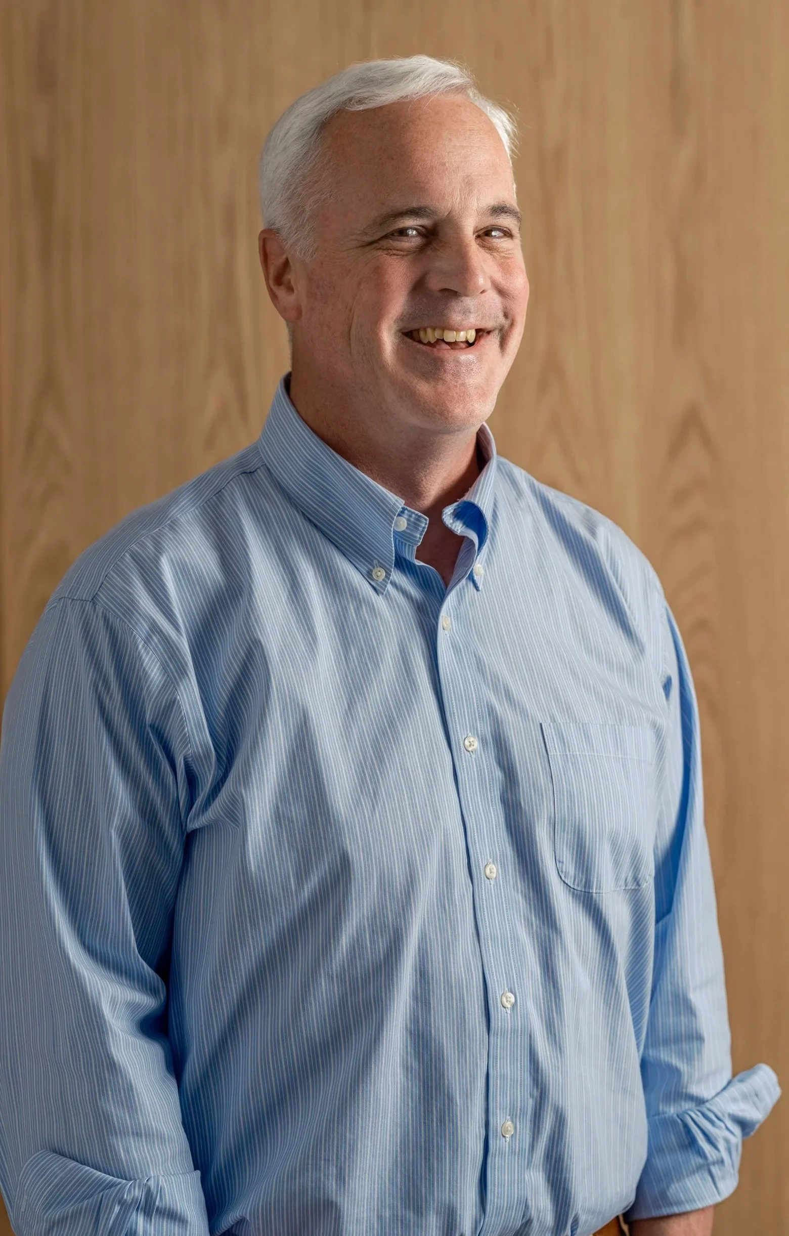 A smiling middle-aged man with gray hair wearing a light blue button-up shirt standing in front of a wooden background.