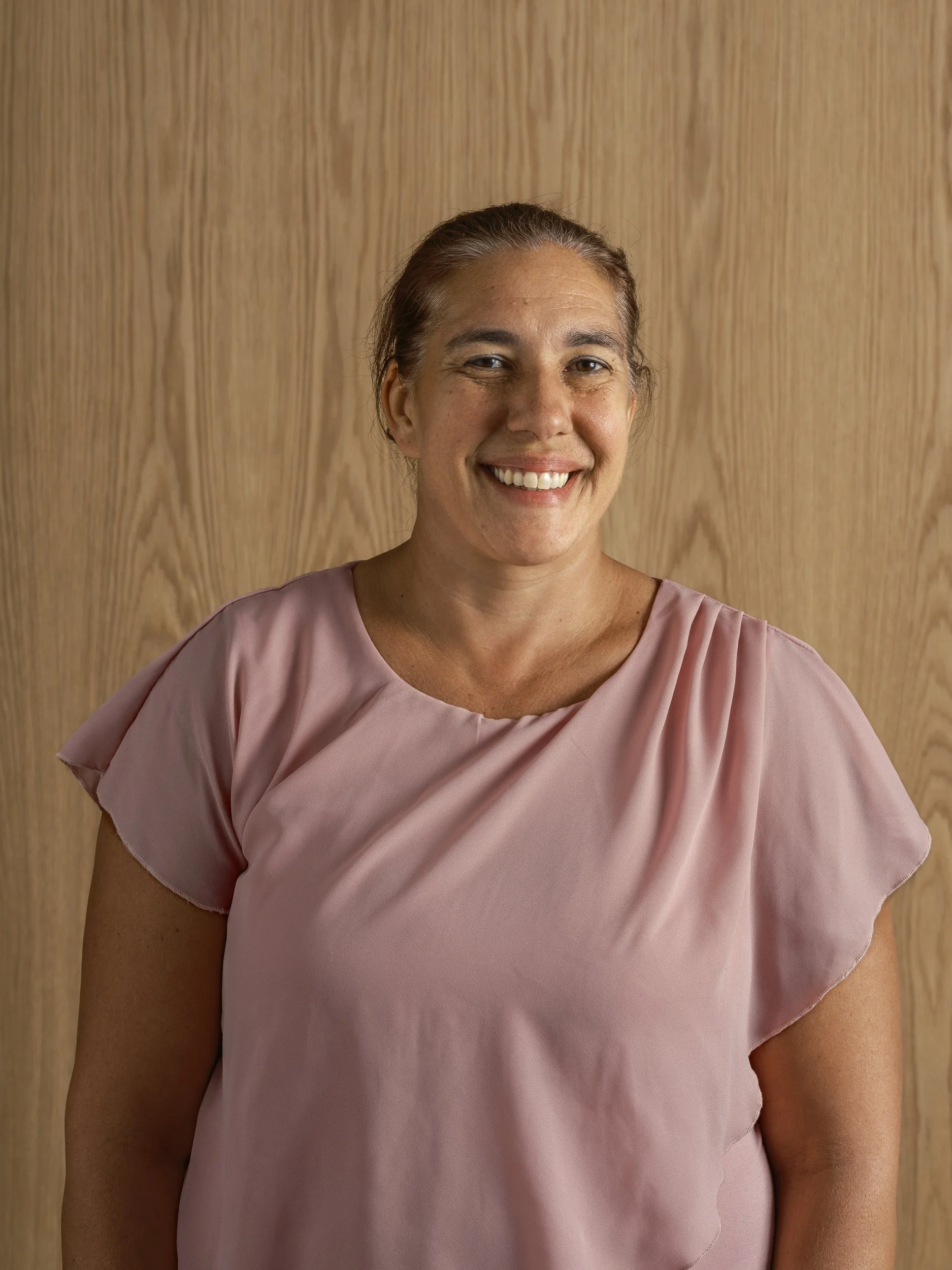 A woman with brown hair tied back, wearing a pink top, standing in front of a wood-paneled background, smiling directly at the camera.