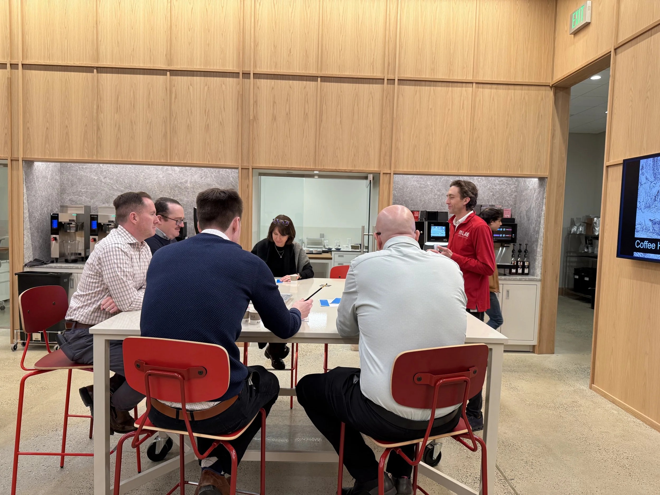 People sitting at a table in a meeting room listening to a man in a red shirt standing and talking.