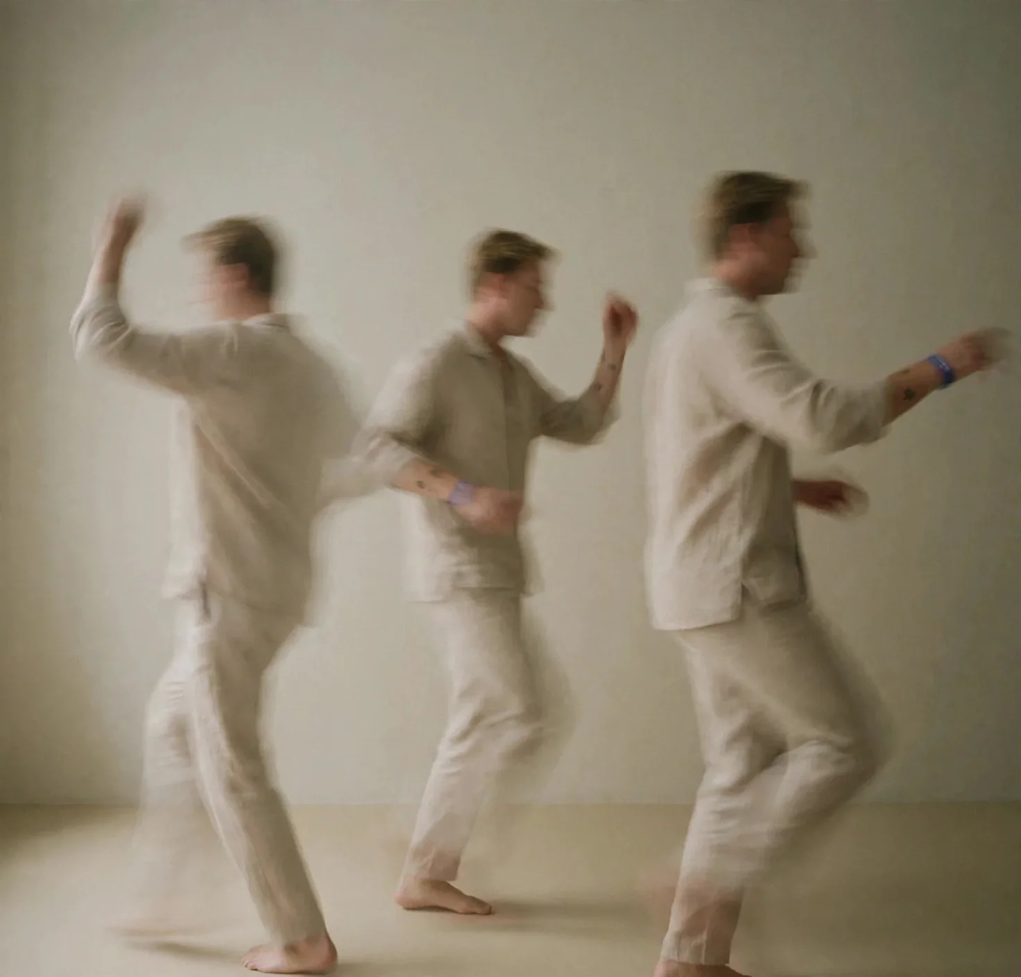 Three people dancing or moving in sequence, blurred, wearing matching beige or light-colored clothing, in an indoor space with plain light-colored wall.