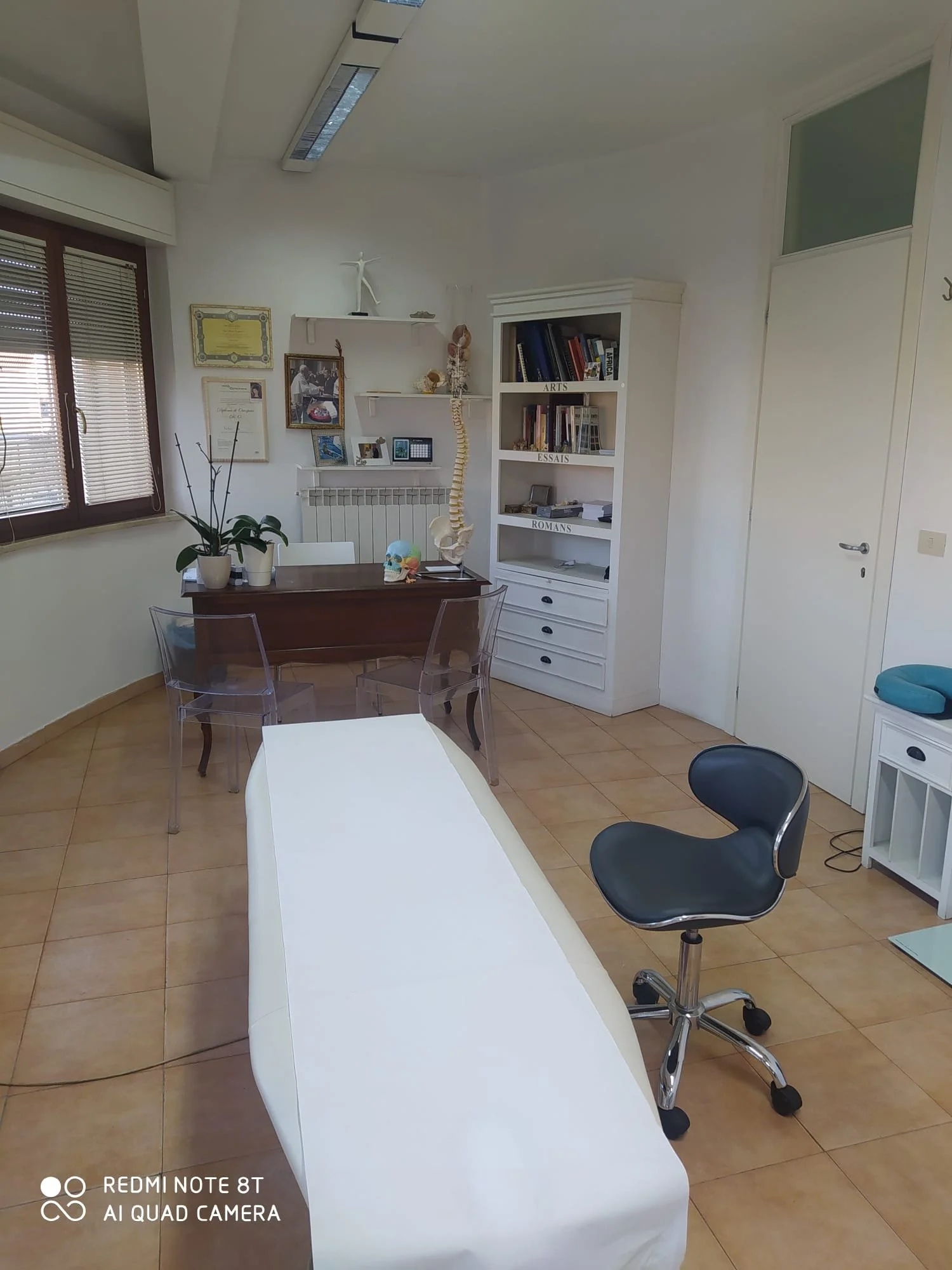 Sala d'ufficio con tavolo bianco, sedia nera, libreria bianca con libri etichettati, decorazioni, rappresentazioni anatomiche, piante verdi, tende alle finestre e illuminazione artificiale.