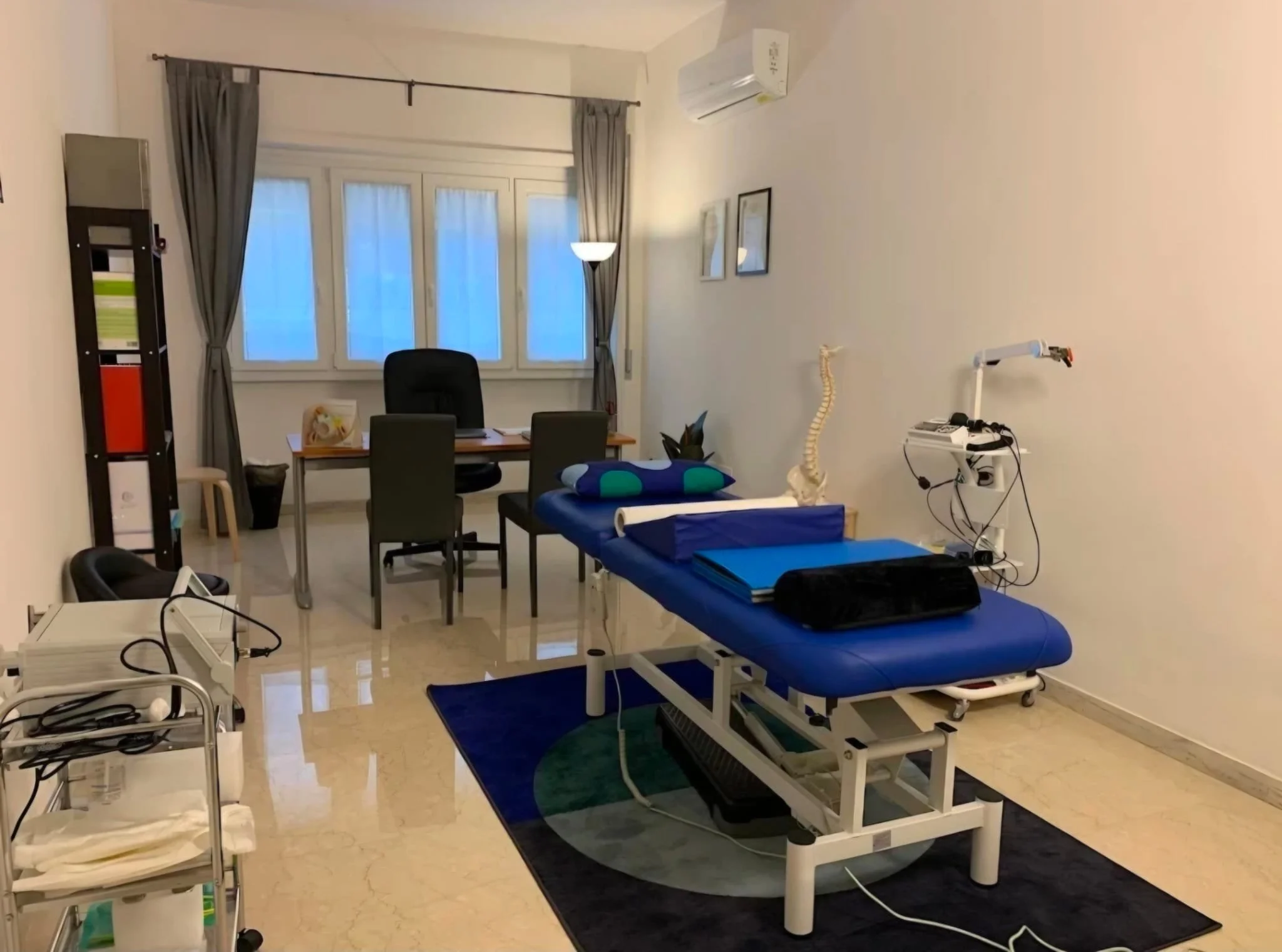 Sala medica con lettino, sedia, apparecchiature mediche e decorazioni semplici su pareti. Ideale per consulti o trattamenti. Grande finestra con tende grigie.