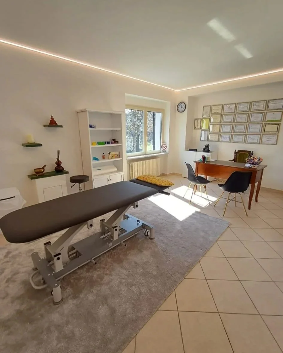 Studio medico o fisioterapia con lettino, librerie, tavolo e sedie, certificati alle pareti e decorazioni.