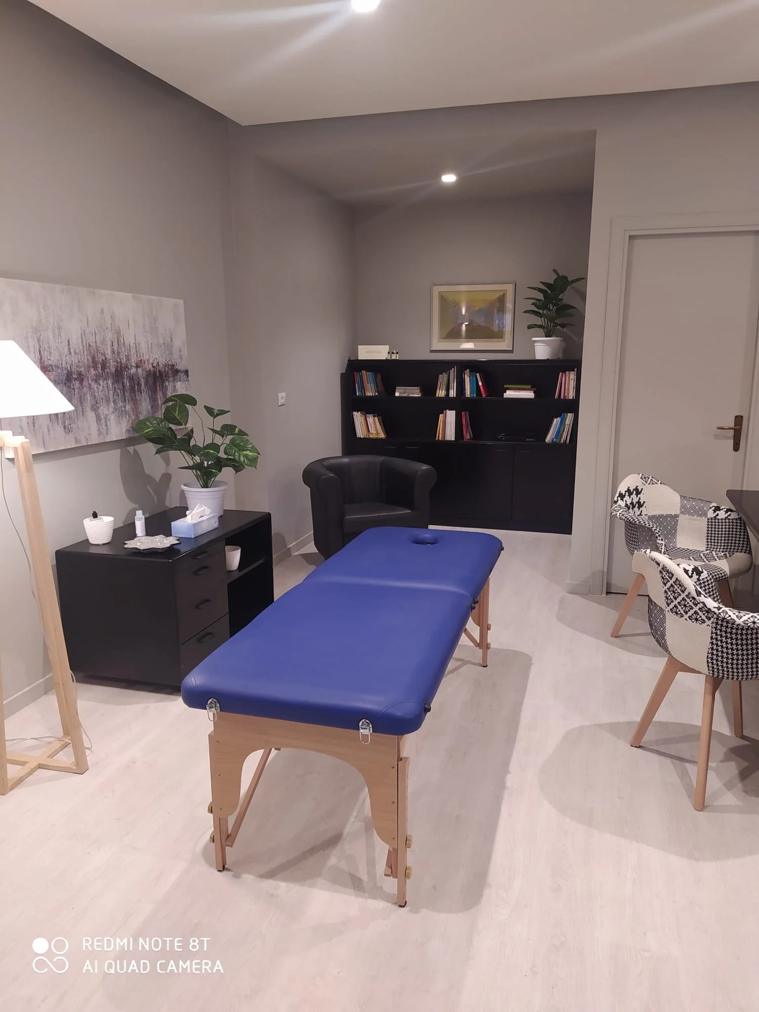 Stanza con massaggiatrice, tavolo da massaggi con rivestimento blu, poltrona nera, libreria con libri e piante, opere d'arte alle pareti, illuminazione soffusa