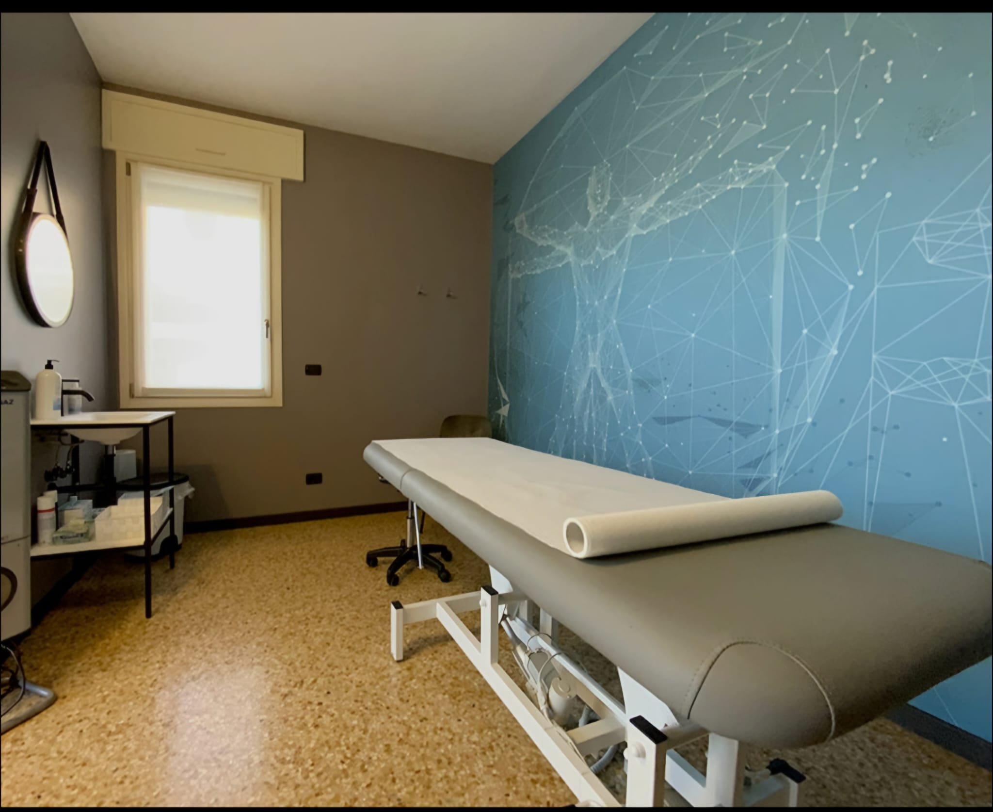 Stanza di medicina con lettino bianco, parete con decorazione digitale a forma di rete di punti e linee, finestra con tende bianche, scaffali con medicazioni e strumenti.