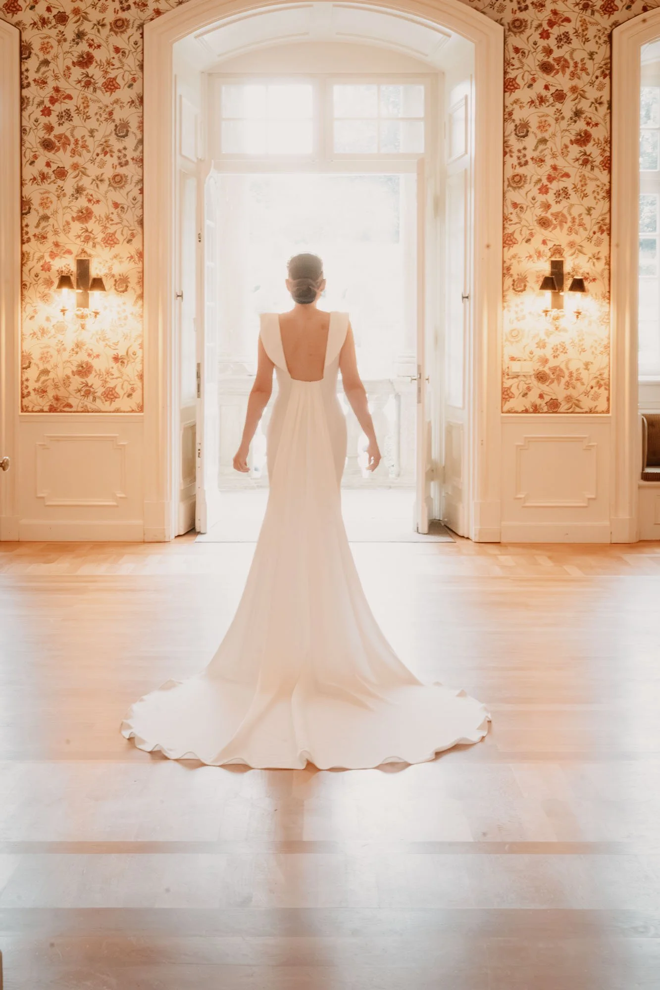 Frau in einem langen weißen Brautkleid steht in einem hellen Raum mit floral tapezierter Wand, große Fenster und helle Holzboden, Blick auf den Rücken der Braut.