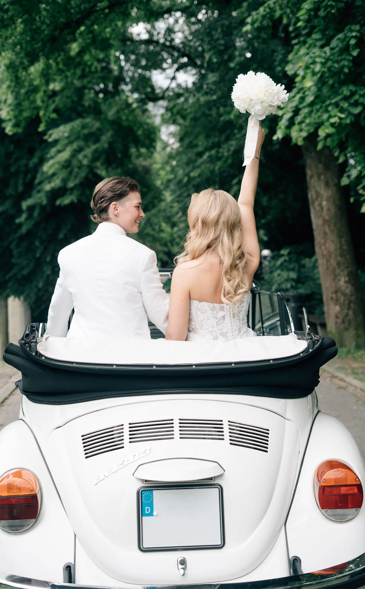 Ein Brautpaar fährt in einem weißen VW Käfer, das Brautkleid ist sichtbar, der Bräutigam trägt einen weißen Anzug, auf einem Weg in einer grünen Parklandschaft, die Braut hält einen weißen Blumenstrauß in der Hand.