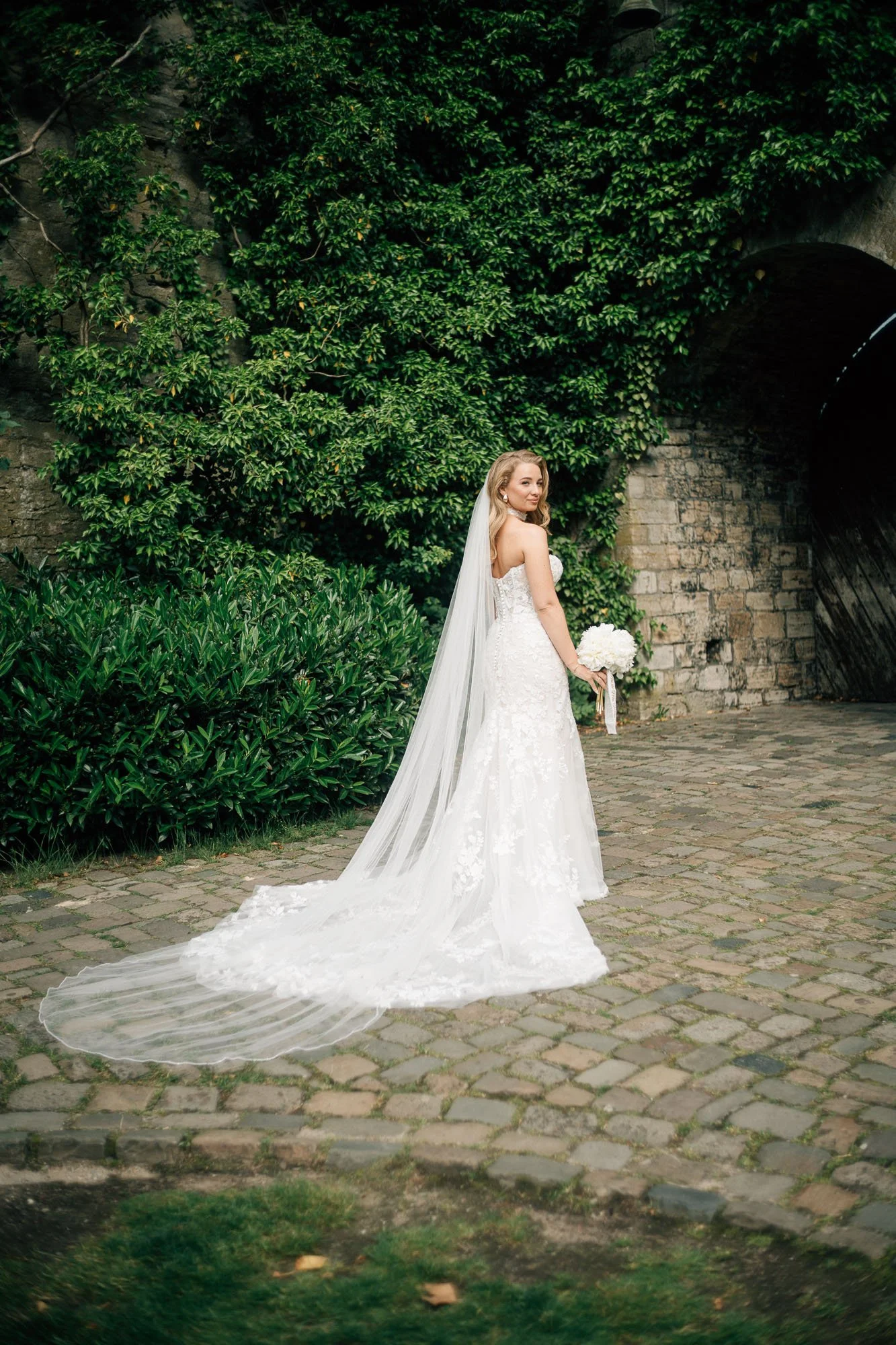 Eine Braut in einem weißen Hochzeitskleid mit Schleier steht vor einer Steinmauer und grünen Pflanzen, hält einen Blumenstrauß in der Hand und lächelt.