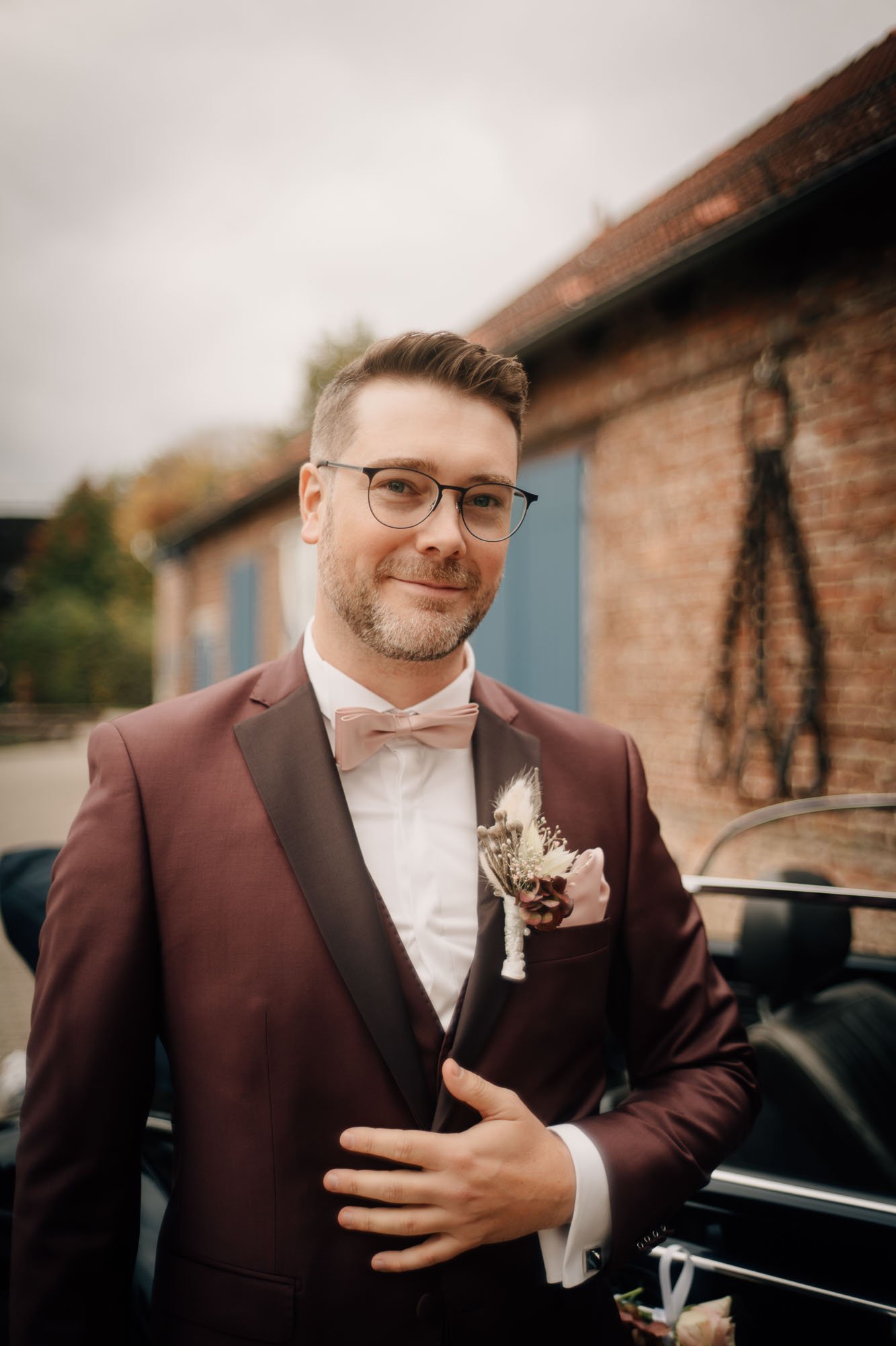 Ein Mann in formeller Kleidung, Brille und mit einem Blumenanstecker am Revers steht neben einem Oldtimer, im Hintergrund eine Ziegelmauer und eine Wolkendecke.