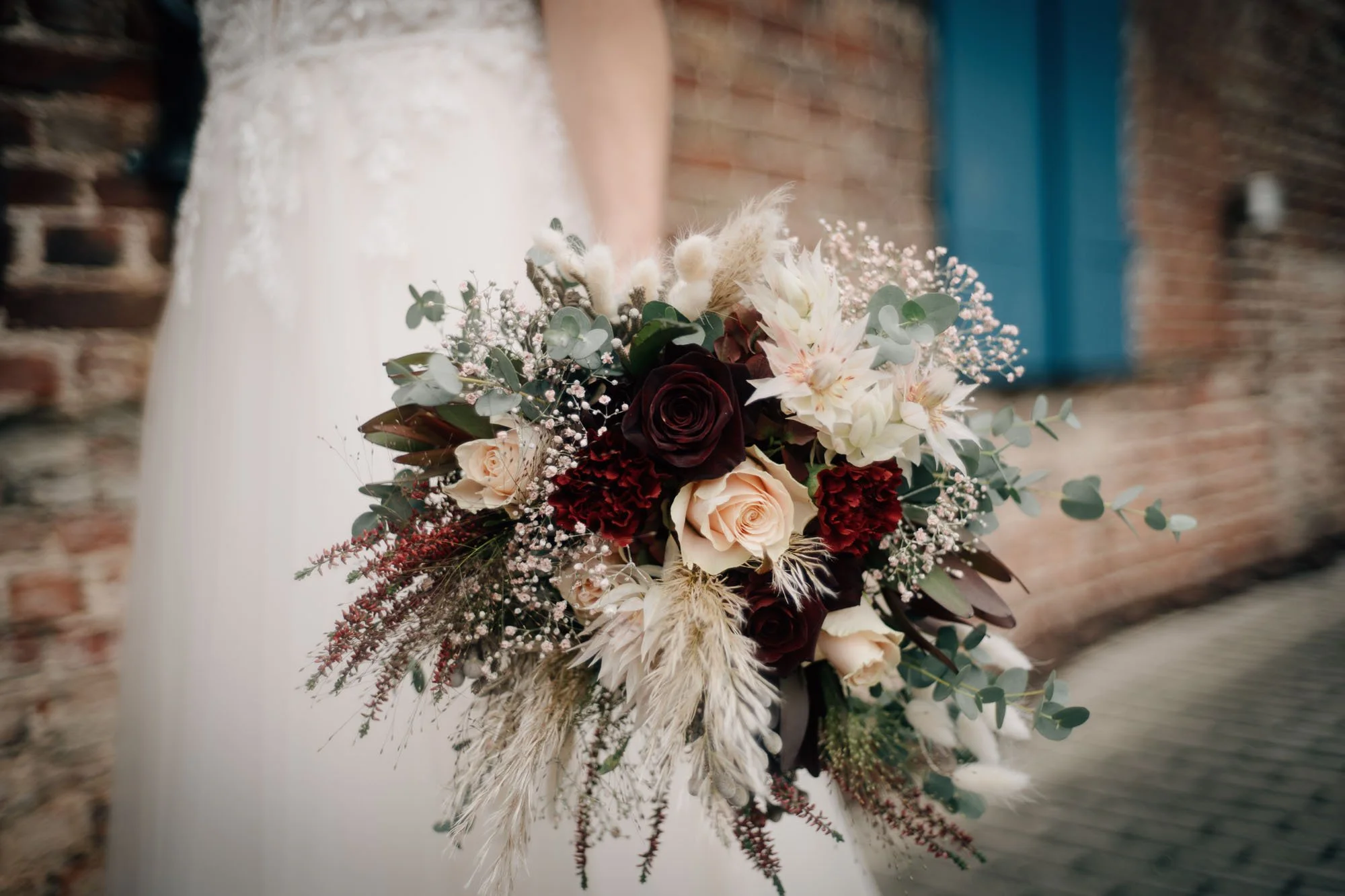 Hochzeitliche Blumenstrauß mit weißen und roten Blumen, Eukalyptus und getrockneten Gräsern vor einer Backsteinmauer.
