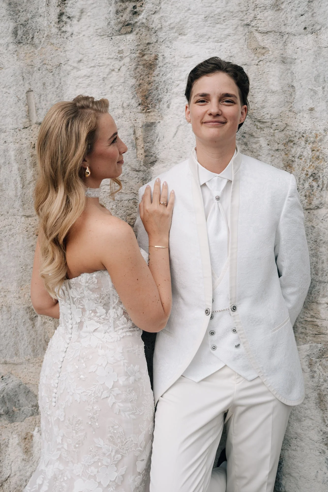 Zwei Frauen in Hochzeitskleidern posieren vor einer alten Steinwand, eine schaut lächelnd die andere an.