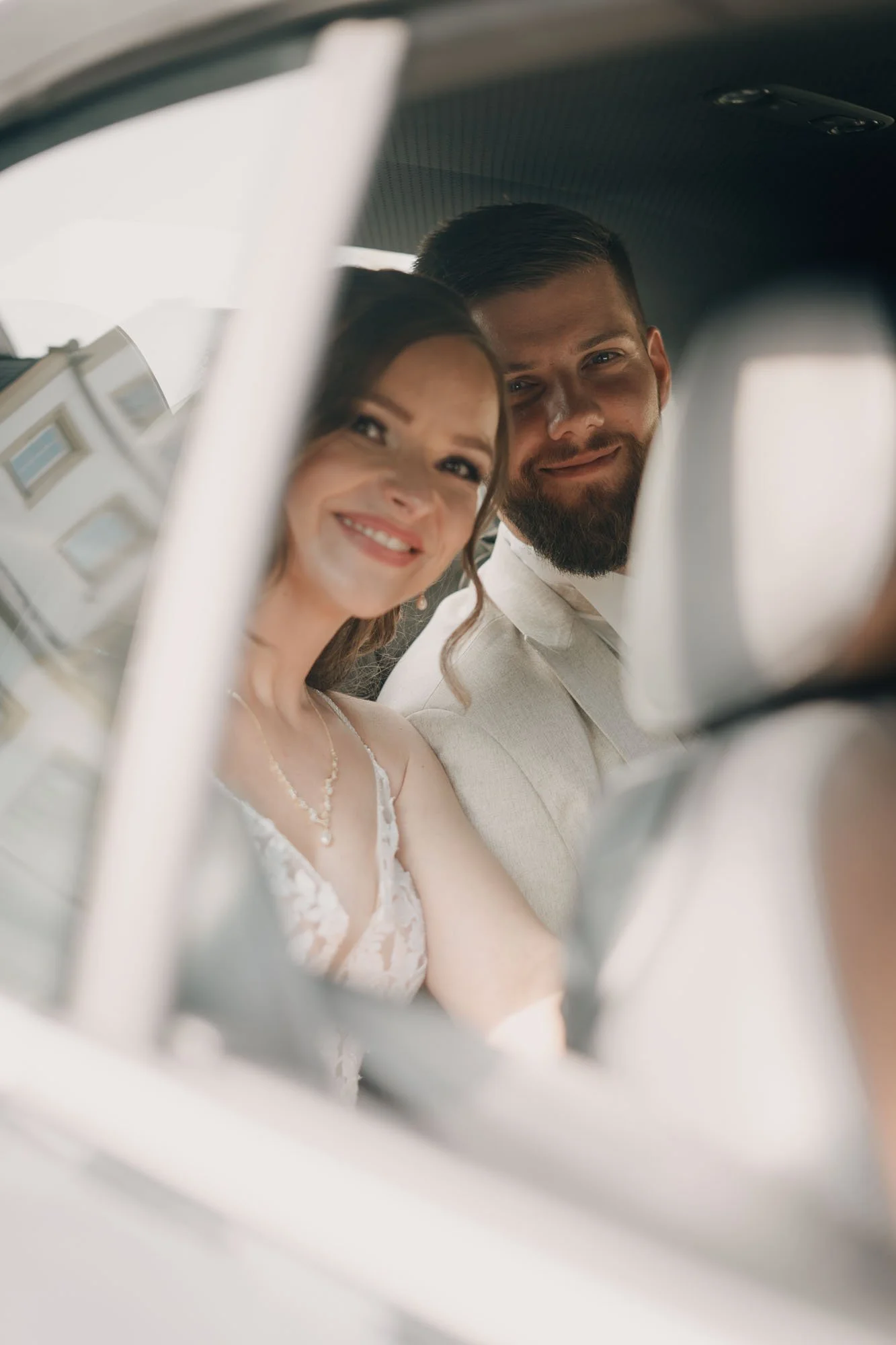 Ein lachendes Brautpaar fotografiert sich in einem Spiegel, aufgenommen durch eine reflektierende Oberfläche, vermutlich im Auto.