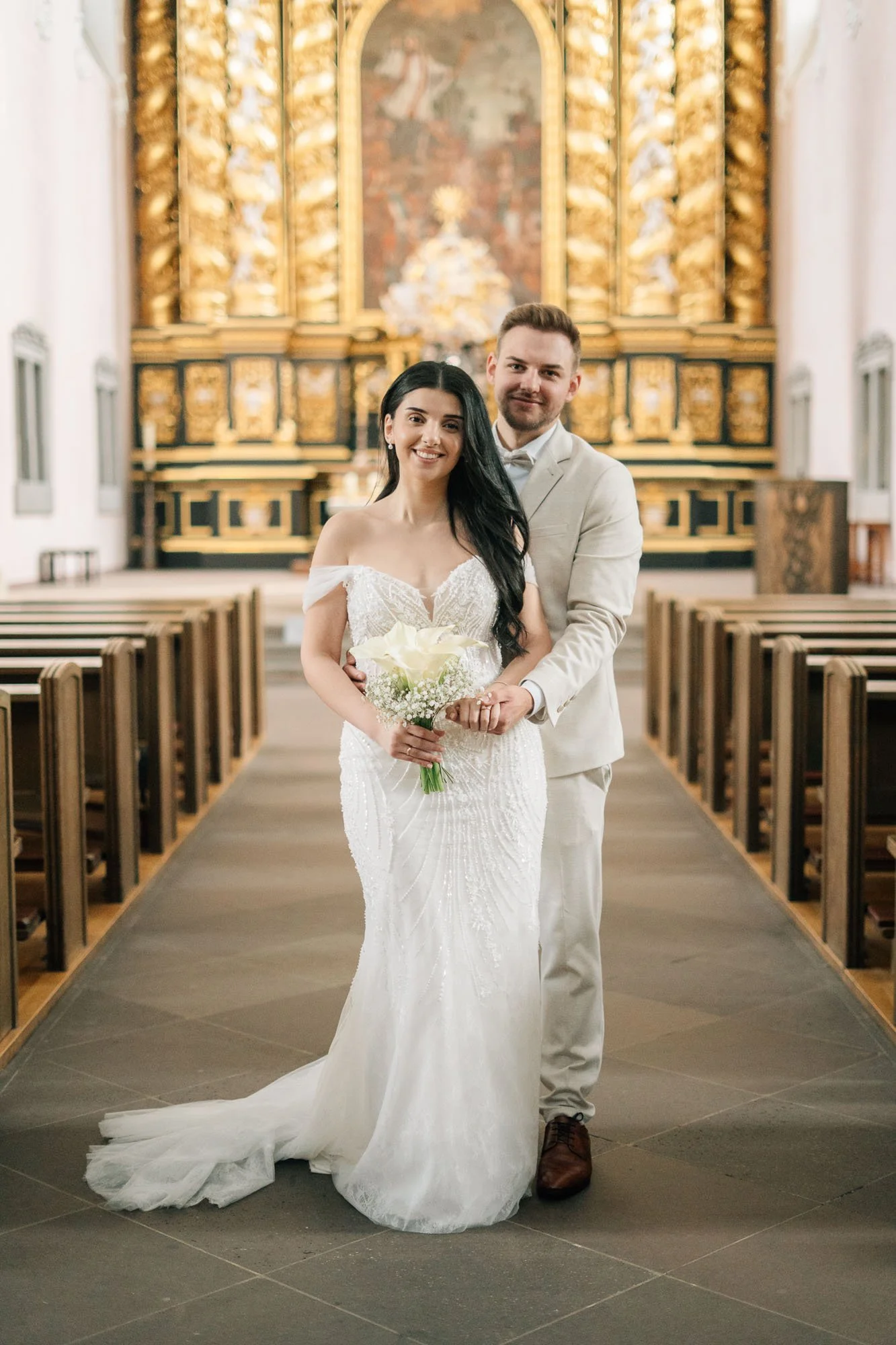 Ein Brautpaar in einer Kirche, die Braut trägt ein weißes Hochzeitskleid und hält einen Blumenstrauß, der Bräutigam trägt einen hellen Anzug. Im Hintergrund befindet sich ein Altar mit Golddekoration und einem Weihnachtsbaum.