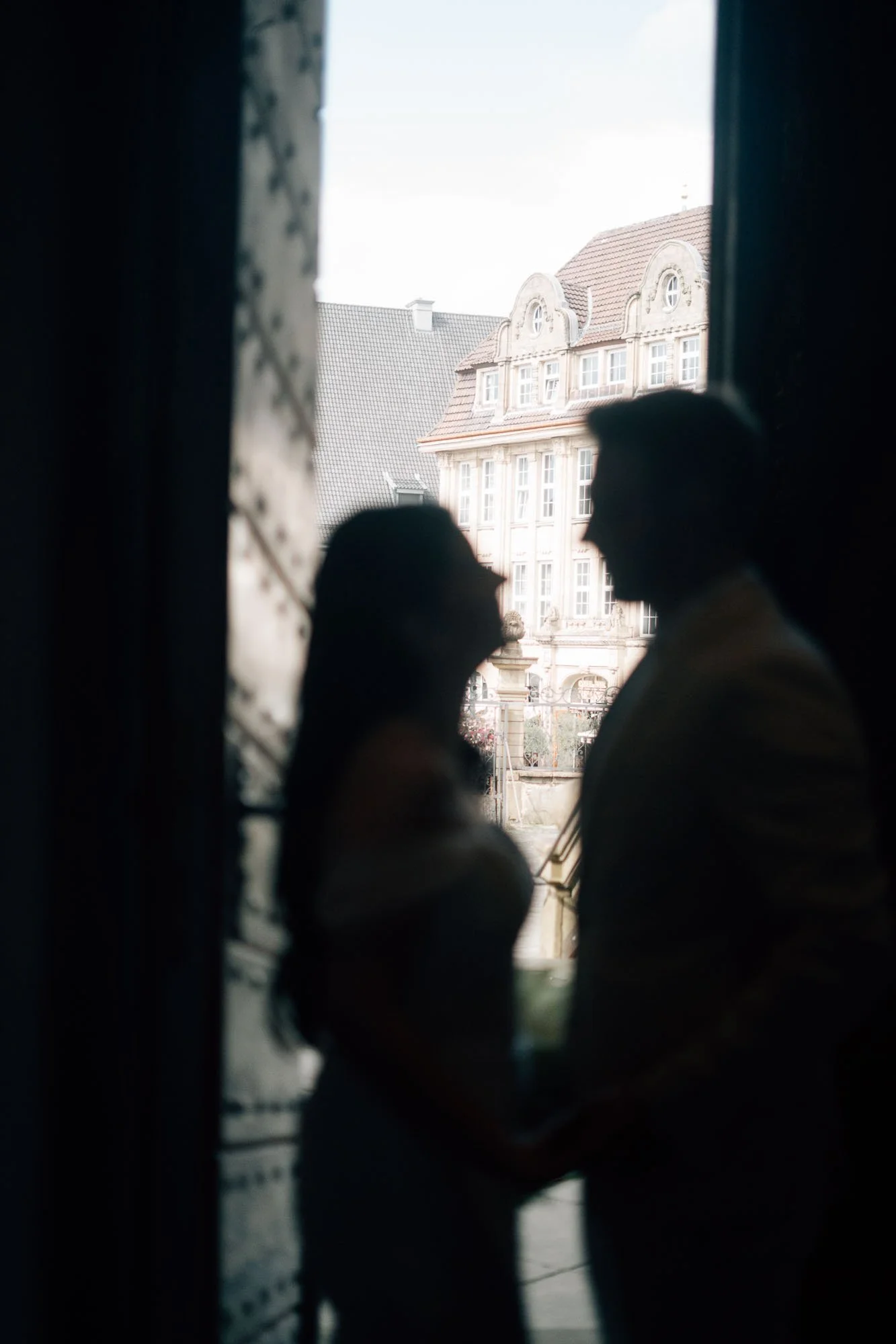 Stilvolle Gebäude mit historischen Fassaden, fotografiert durch ein Fenster mit Vorhängen, im Vordergrund sind unscharfe Silhouetten von Menschen.