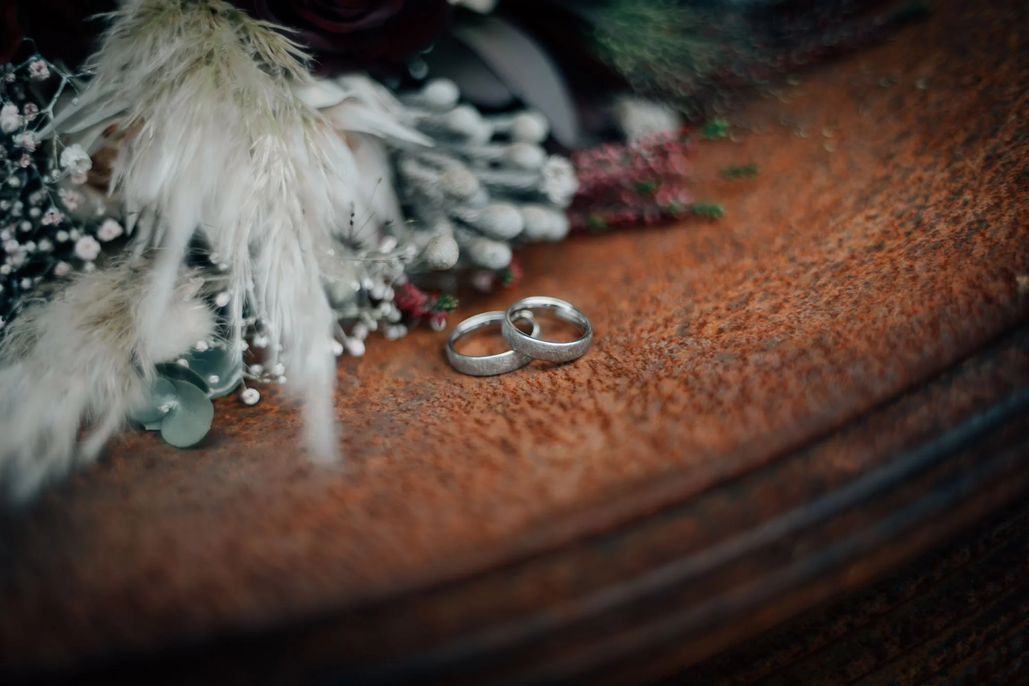 Silberne Eheringe liegen auf einem rustfarbenen Steinboden, im Hintergrund ist eine Hochzeitdekoration mit weißen und pinken Blumen und Federn sichtbar.