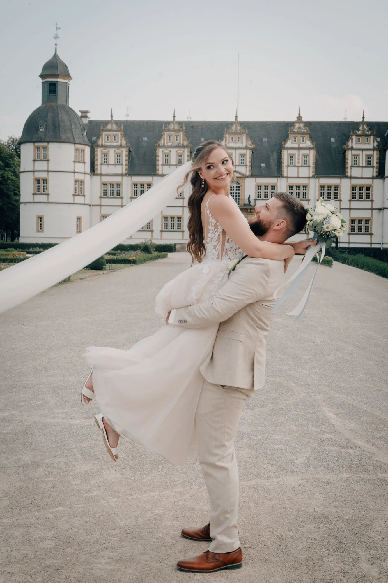 Ein verliebtes Paar an einem historischen Schloss im Freien, vermutlich bei einer Hochzeit, mit dem Mann, der die Frau hochhebt, beide lächelnd.