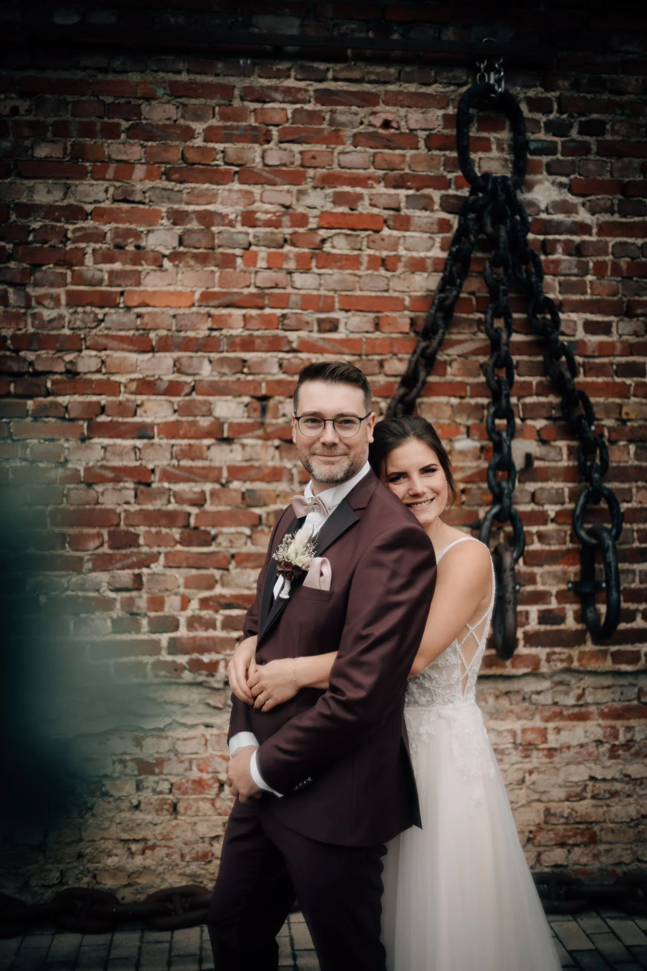 Ein Paar in Hochzeitskleidung posiert vor einer roten Backsteinmauer. Der Mann trägt einen braunen Anzug mit Fliege, die Frau trägt ein weißes Brautkleid.