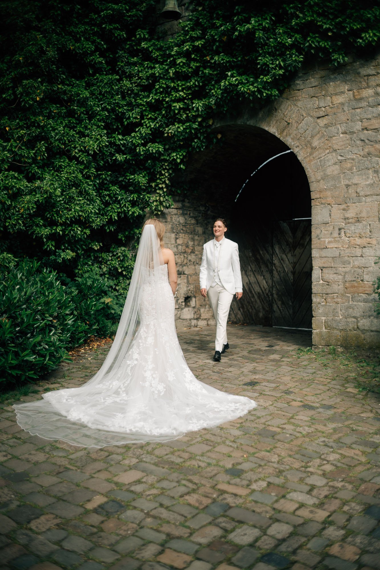 Ein Brautpaar, die Braut trägt ein weißes Hochzeitskleid mit Schleier und der Bräutigam trägt einen weißen Anzug, stehen sich gegenüber vor einer alten Steinmauer mit einem Tor und umgeben von grünen Pflanzen auf einem gepflasterten Boden.