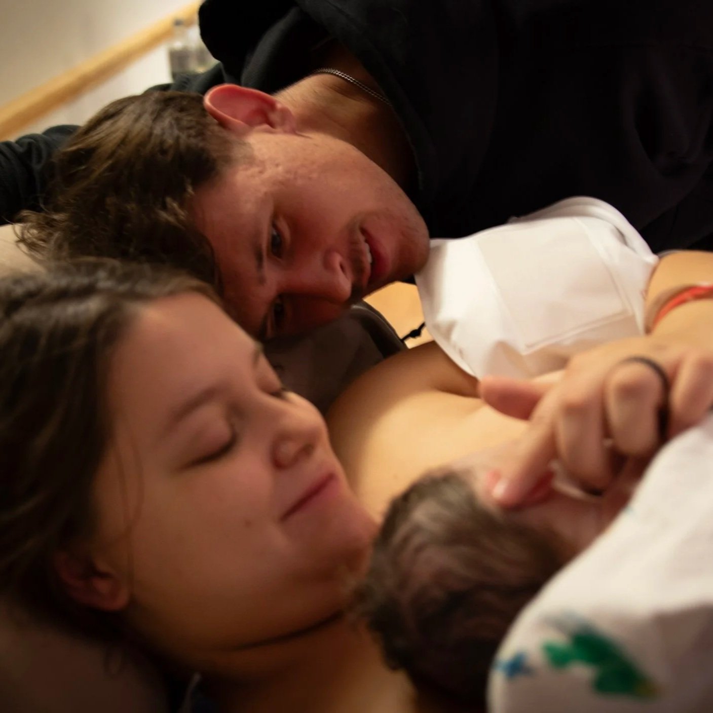 Two people, a man and a woman, lying close together with their eyes closed, and a newborn baby in a hospital room.