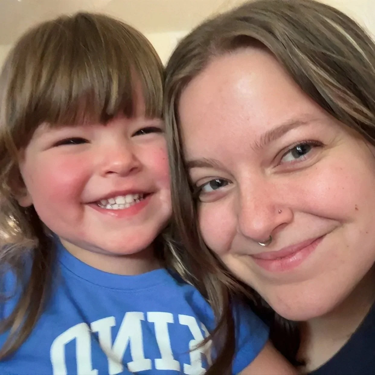 Close-up of a smiling woman with brown hair, light skin, and a nose piercing, taking a selfie with a young girl with brown hair, light skin, and a big smile showing teeth.