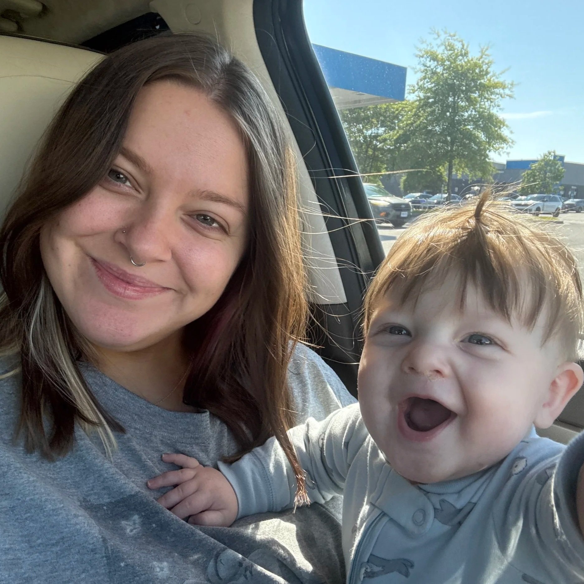 A woman with brown hair smiling and a young boy with blonde hair smiling and with mouth open in a car, with a parking lot and trees outside.