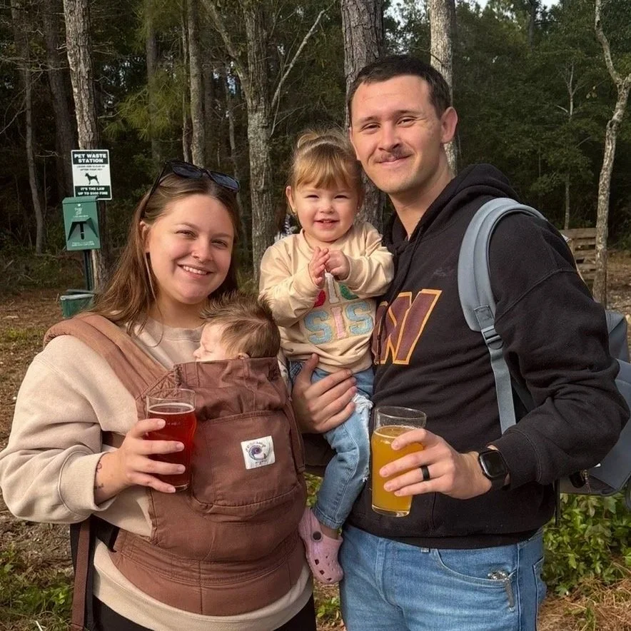 A family of four outdoors near a wooded area, with a woman holding a baby in a front carrier, a man holding a young girl, both with drinks.