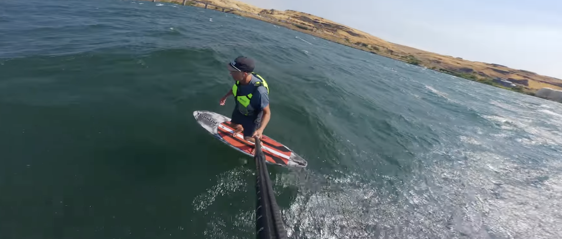 A person riding a hydrofoil down winding  on a body of water, holding a selfie stick, with a shoreline in the background.