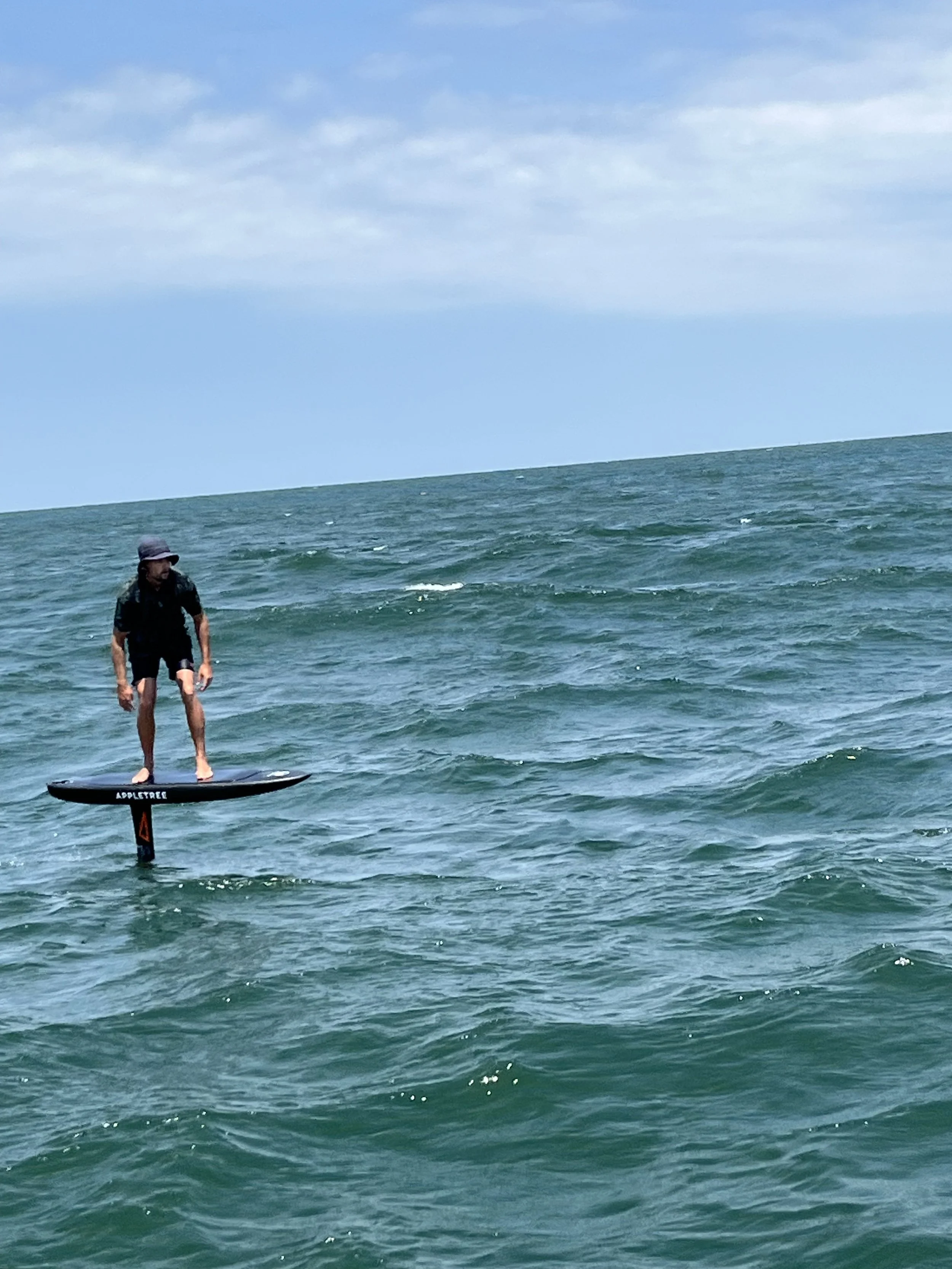 A person standing on a SUP foil board down winding in the ocean, wearing a hat and dark clothing, with cloudy skies above.