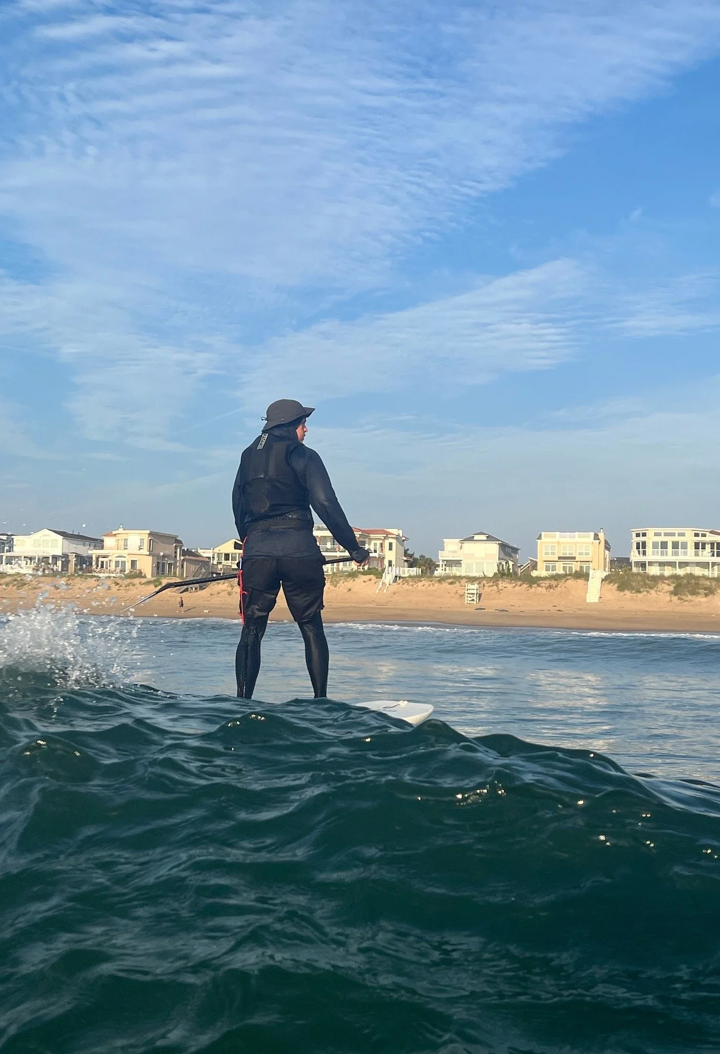 Person wearing a black wetsuit and hat Stand Up Hydrofoil on the ocean near a sandy beach with houses in the background under a cloudy sky.