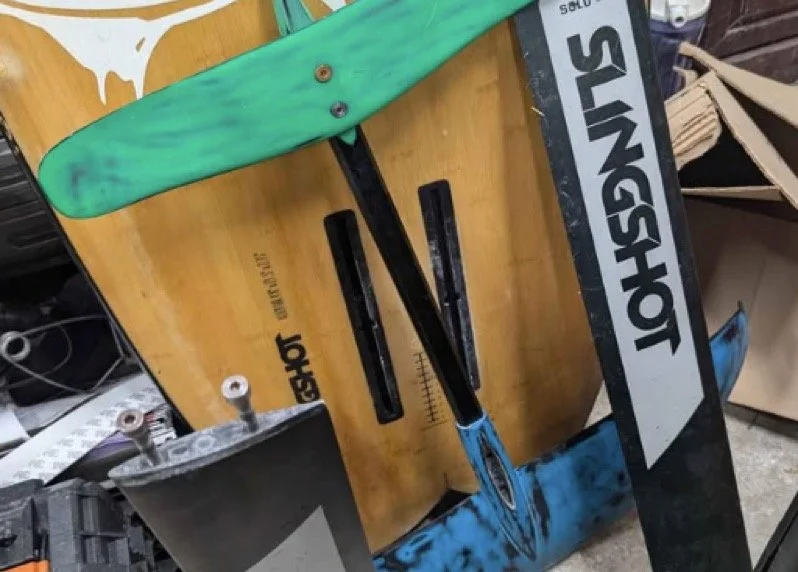A hydrofoil and a pair of boots on a wooden surface next to a snowboard, with some electronic equipment and cables in the background.