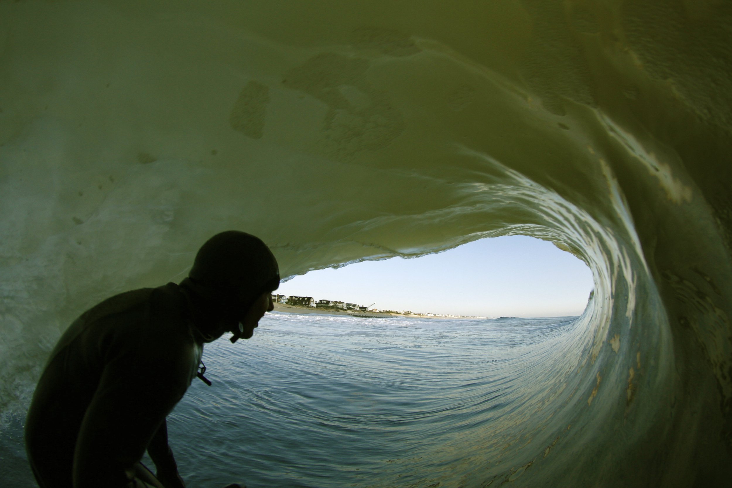 Inside a wave with a person silhouetted at the front, getting tubed or barreled overlooking the ocean and a coastline with buildings in the distance.