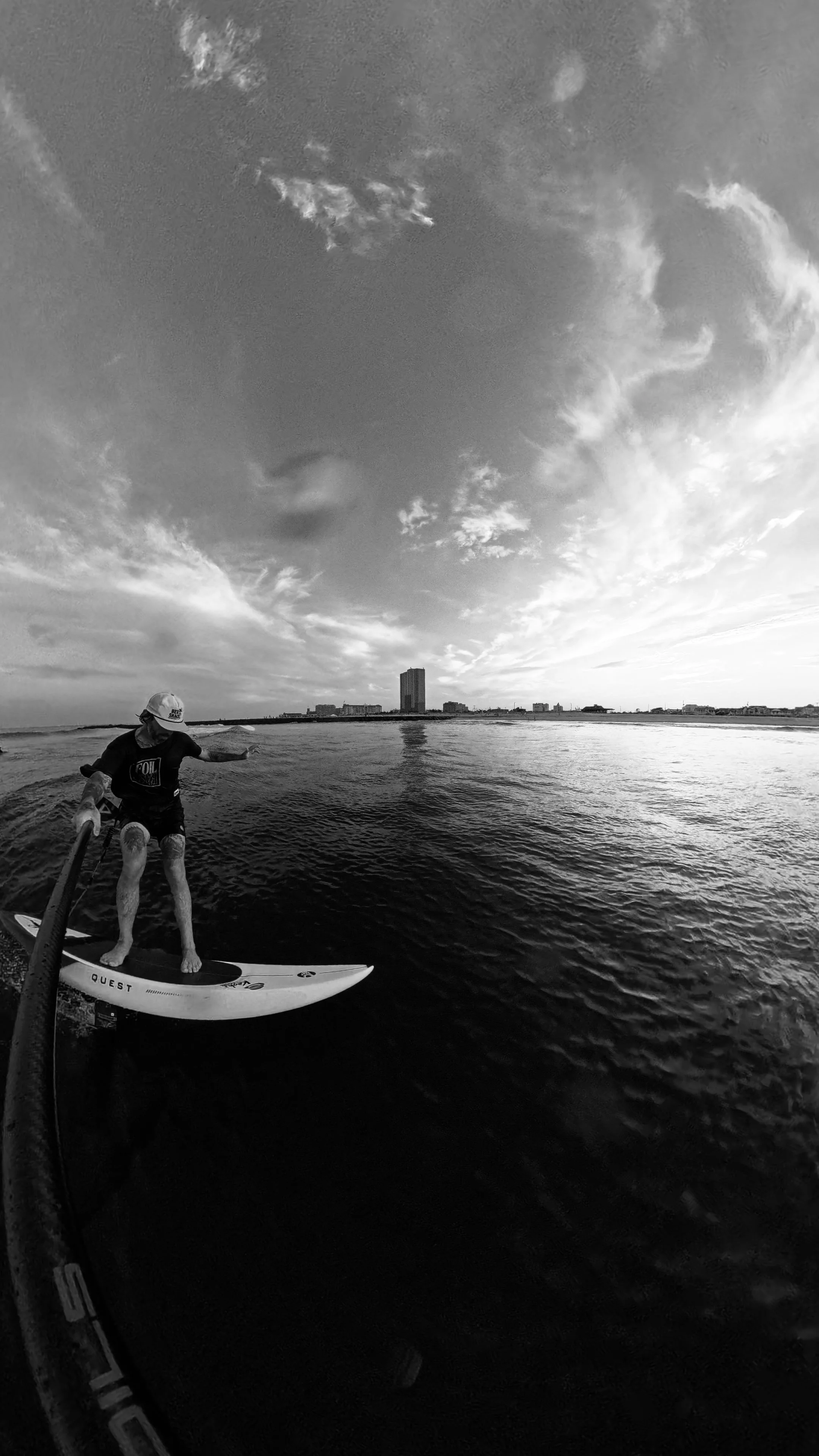 A person hydro foiling on a Stand Up Paddleboard on calm water, wearing a baseball cap and shorts, with a city skyline in the background under a partly cloudy sky.
