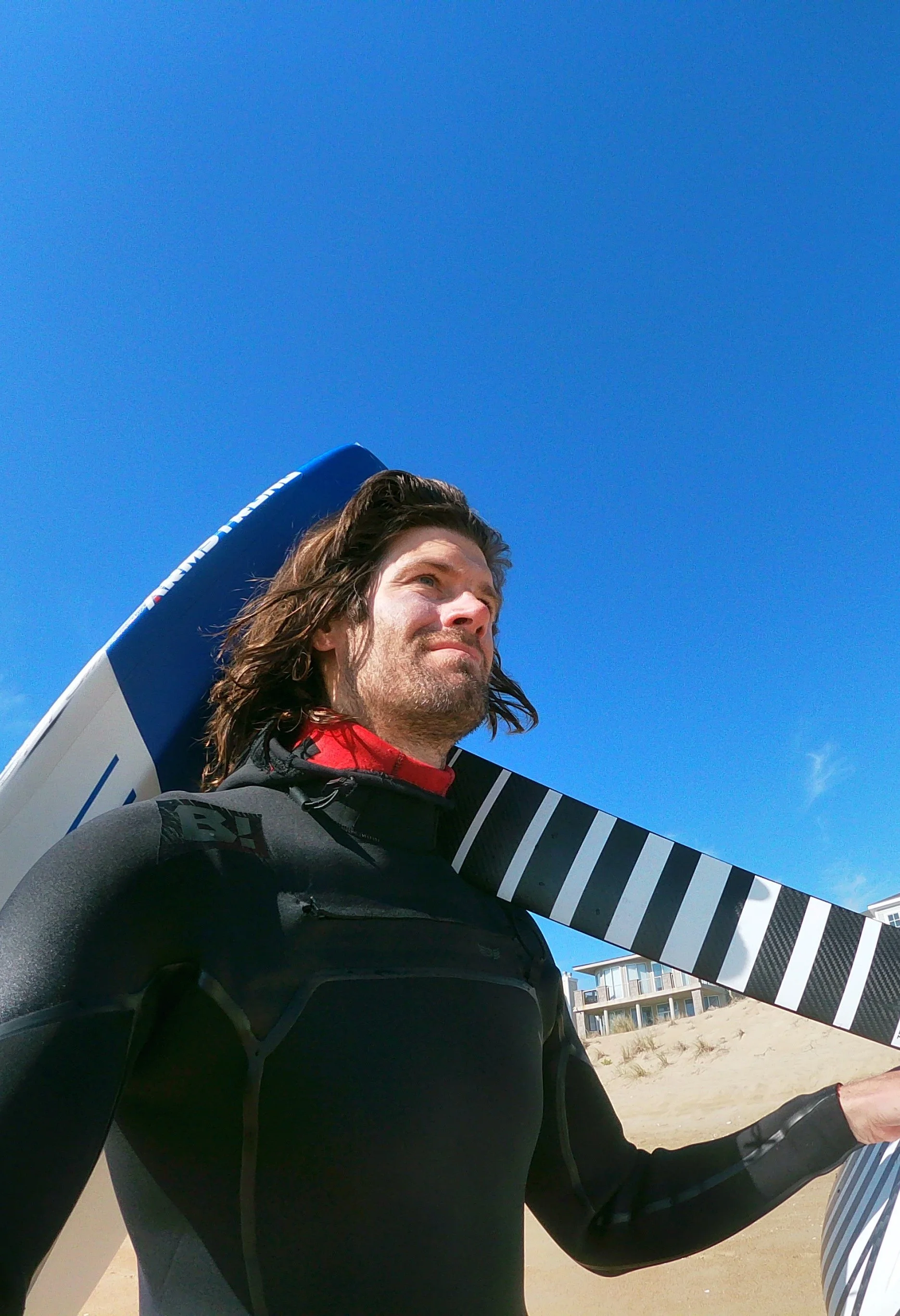 A man with long hair wearing a black wetsuit, standing on a beach, carrying a hydrofoil by Armstrong foils.