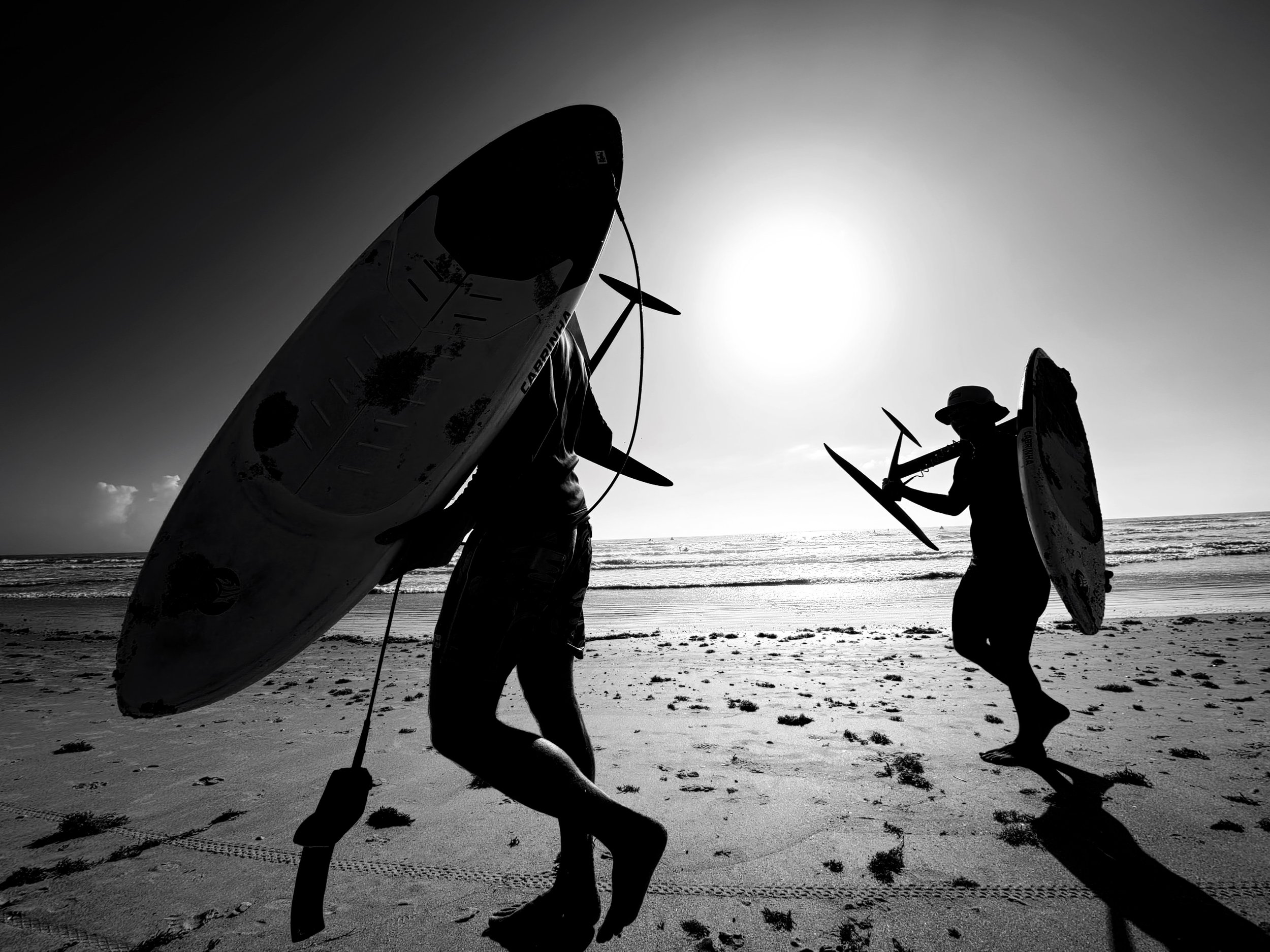 Silhouettes of three people carrying surfboards on a beach during sunset or sunrise, with visible footprints and a smooth horizon in the background.